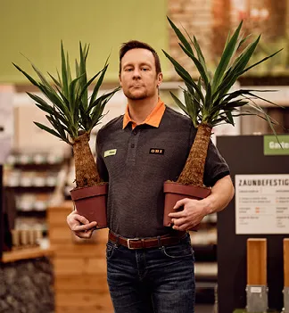 Mitarbeiter von OBI AT GmbH im Markt, trägt ein graues Polo mit OBI-Logo, steht vor einer "GartenPlaner"-Tafel.