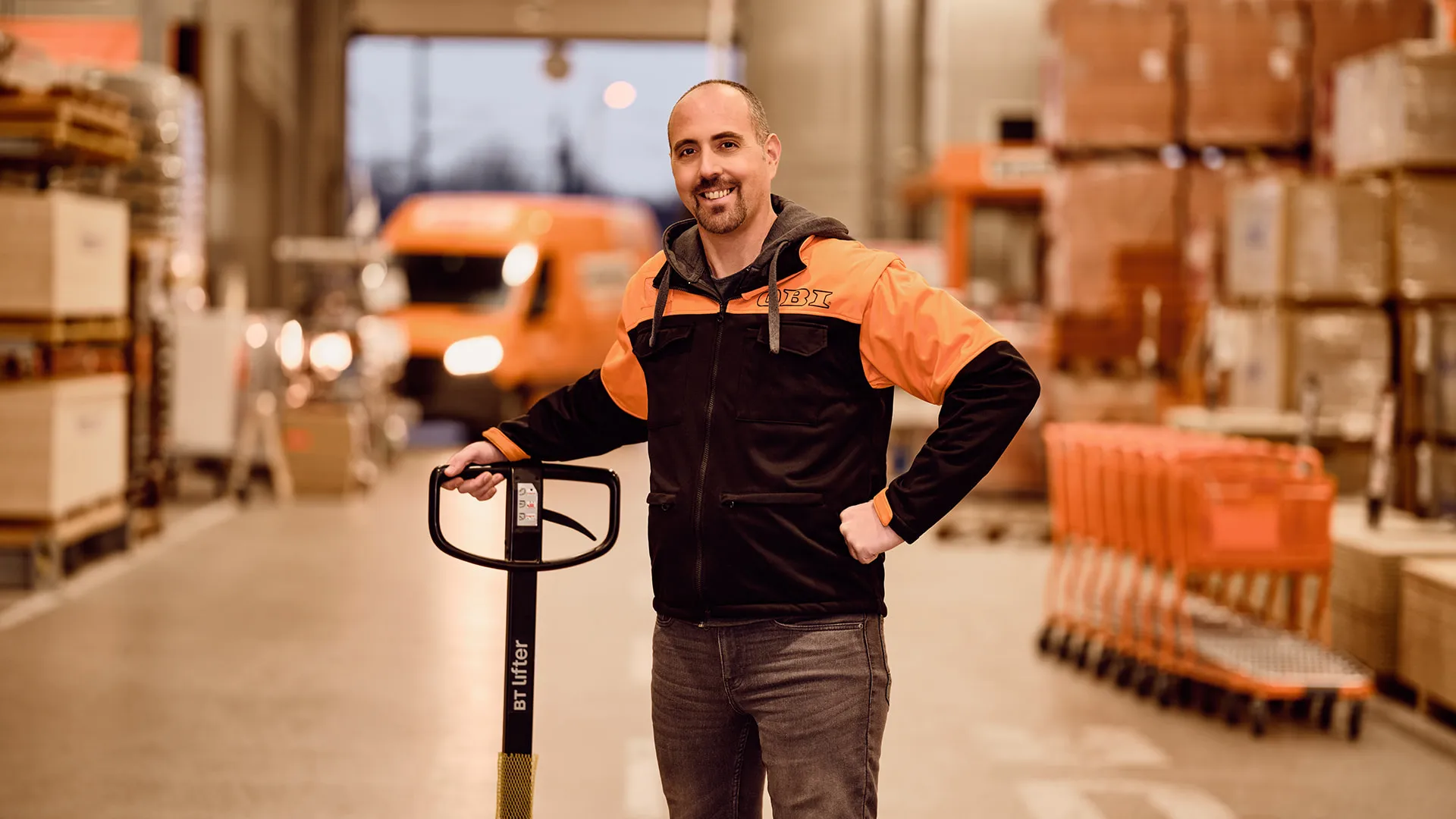 Ein Mitarbeiter von OBI AT GmbH steht in einem Lager, lächelt und hält einen Hubwagen.
