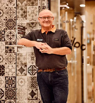 Ein Mitarbeiter von OBI AT GmbH in einem Baumarkt, lächelnd, in der Badabteilung mit einem Laptop und Plänen auf dem Tisch.