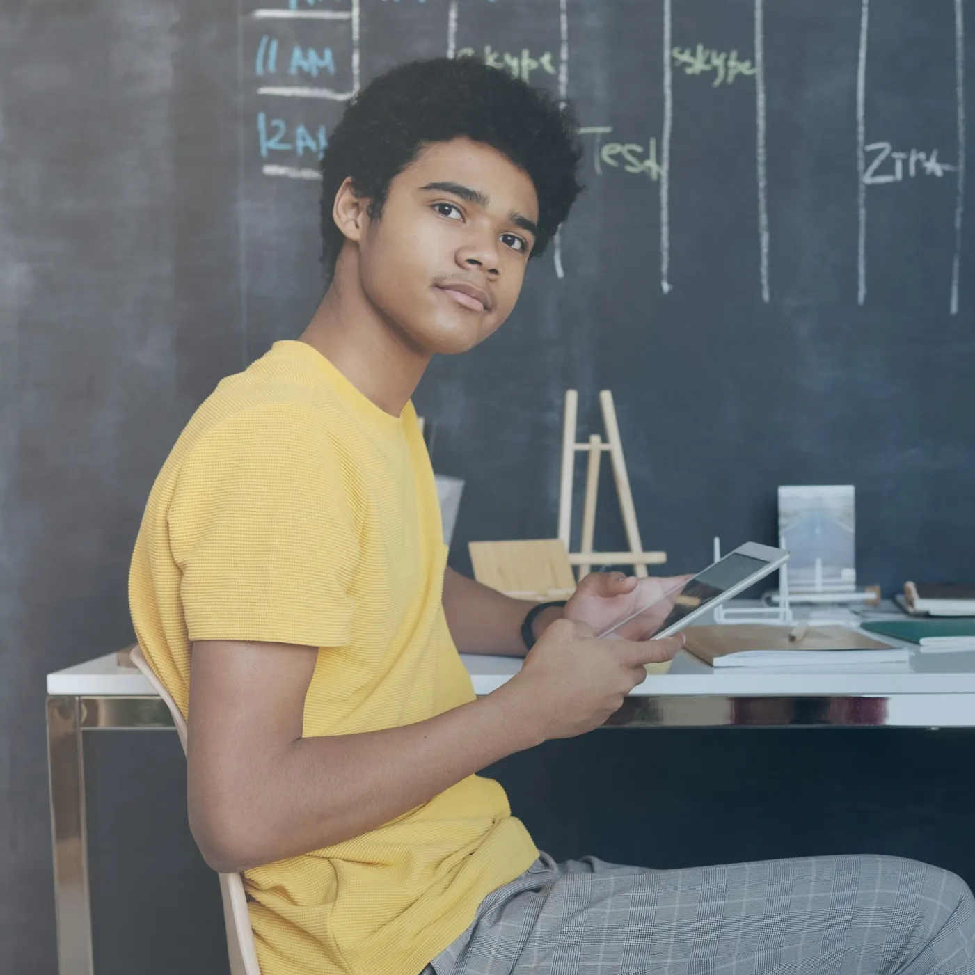 Ein junger Mann in einem gelben T-Shirt sitzt an einem Schreibtisch und hält ein Tablet, im Hintergrund eine Tafel mit Notizen.