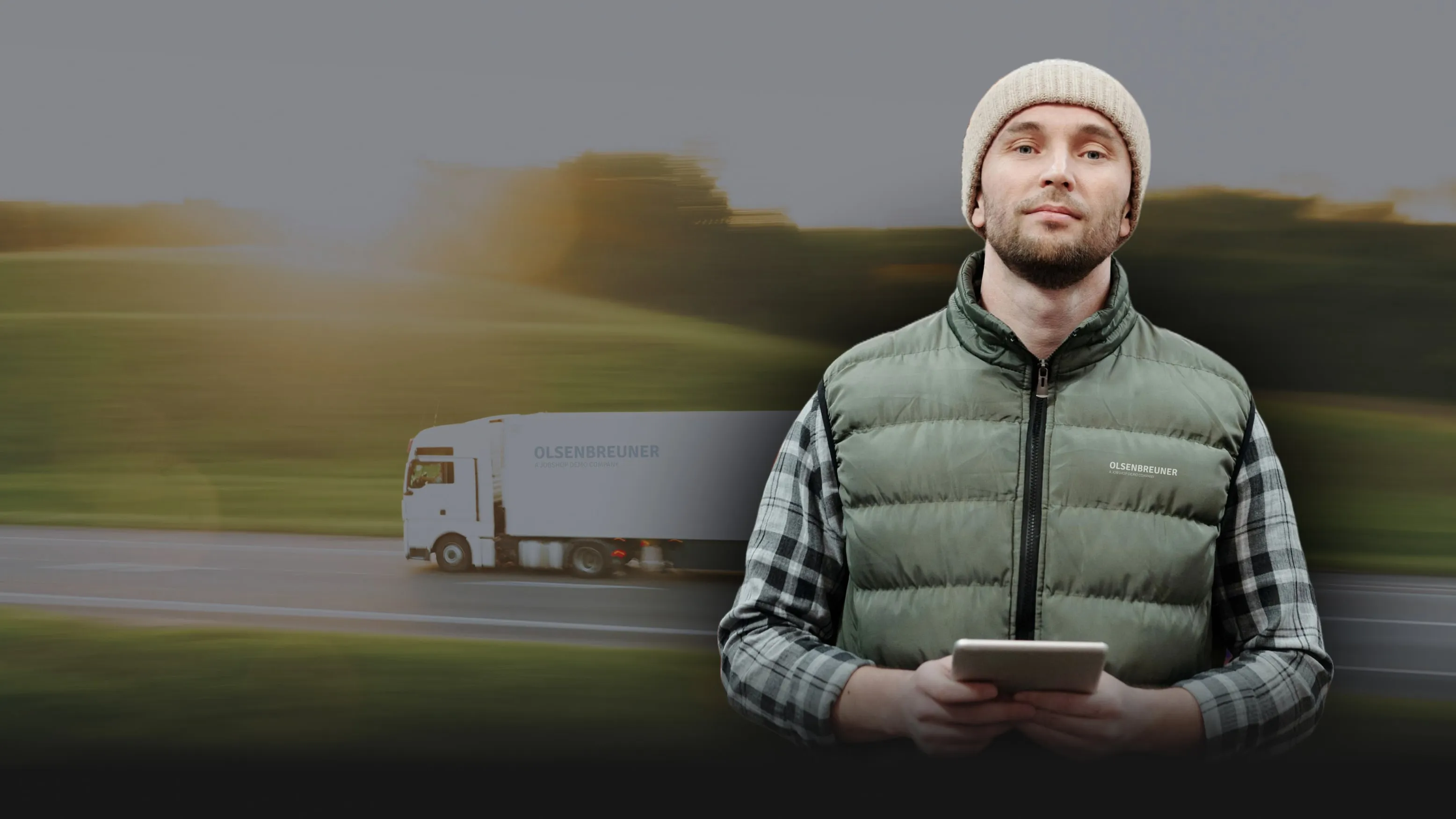 Ein Mann mit Mütze und Tablet steht vor einem vorbeifahrenden Olsen & Breuner LKW.