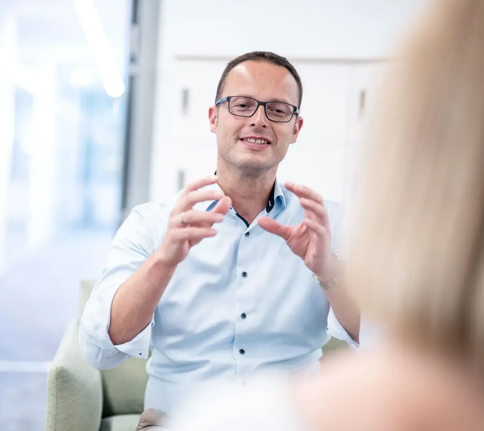 Ein Mann in einem Gespräch, trägt ein hellblaues Hemd und eine Brille. Er gestikuliert, um einen Punkt zu verdeutlichen.
