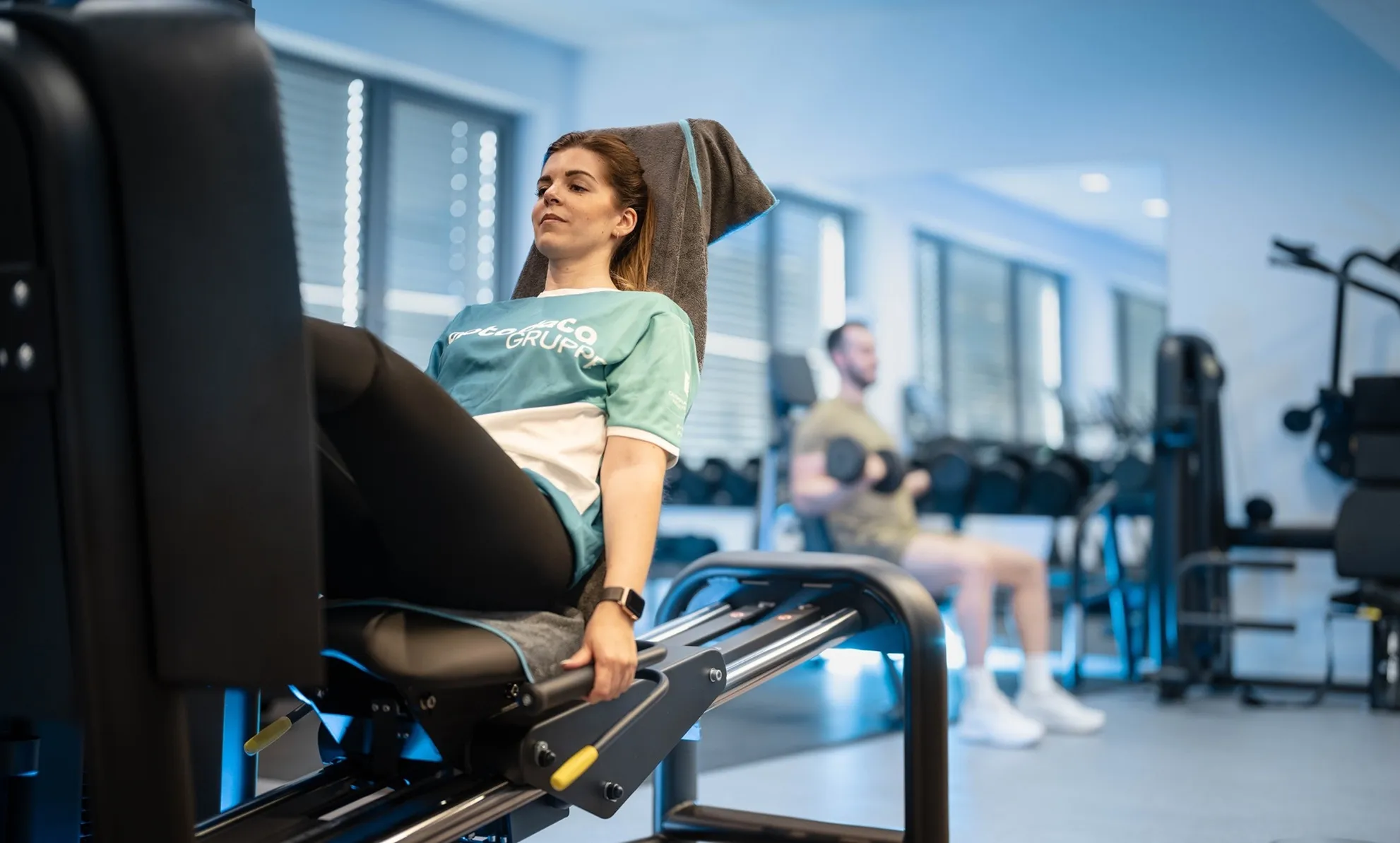 Ein Mitarbeiter der opta data Gruppe trainiert im Fitnessstudio, während eine Frau an einer Beinpresse trainiert.
