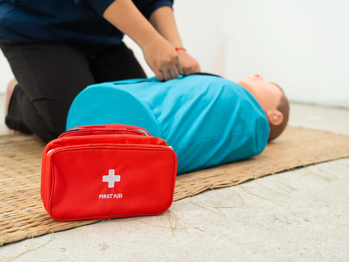 Eine Person übt an einer Puppe Erste-Hilfe-Maßnahmen. Im Vordergrund liegt eine rote Erste-Hilfe-Tasche.