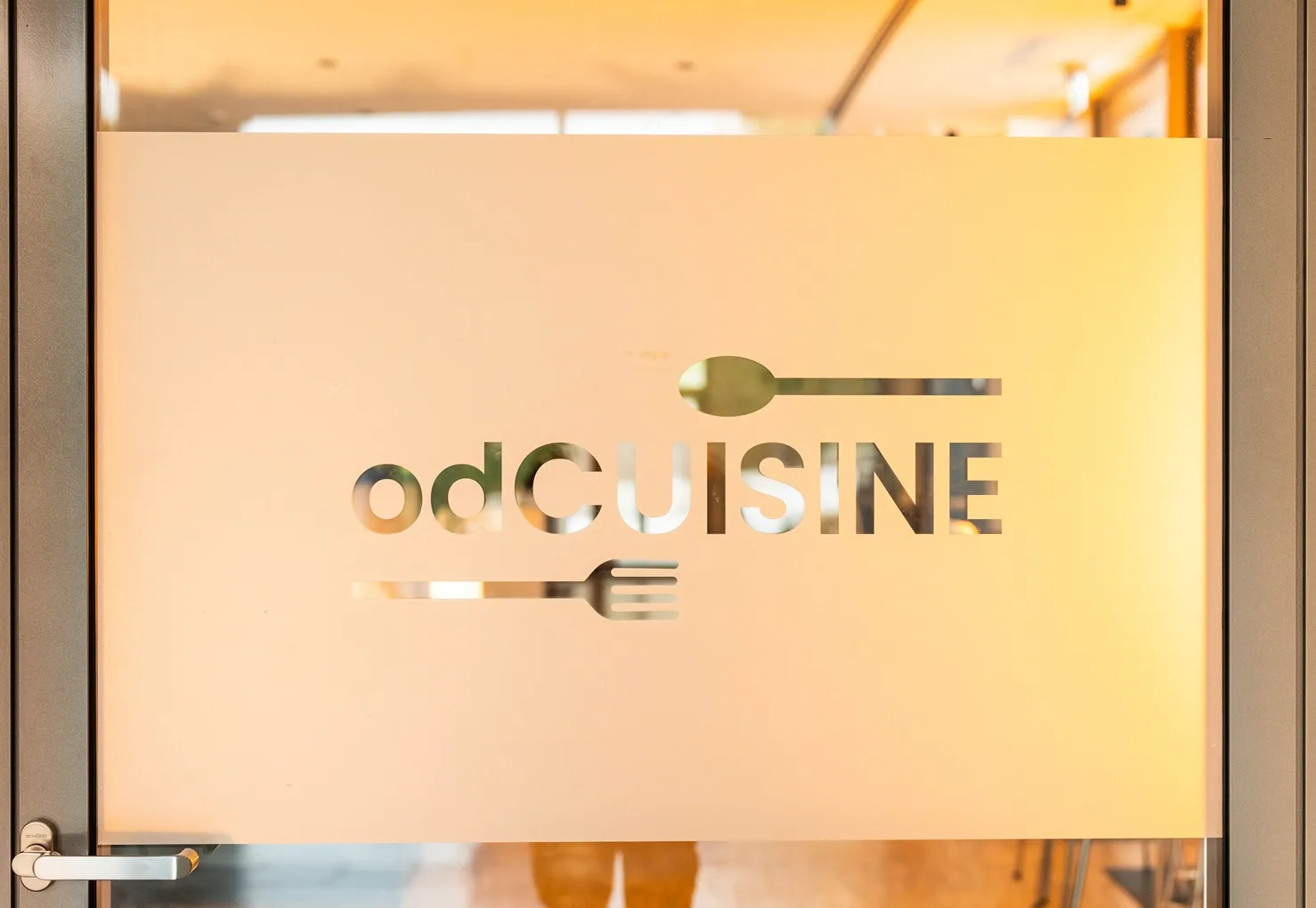 Glaswand mit dem Schriftzug 'odCUISINE' und Symbolen von Löffel und Gabel in einem modernen Bürogebäude.
