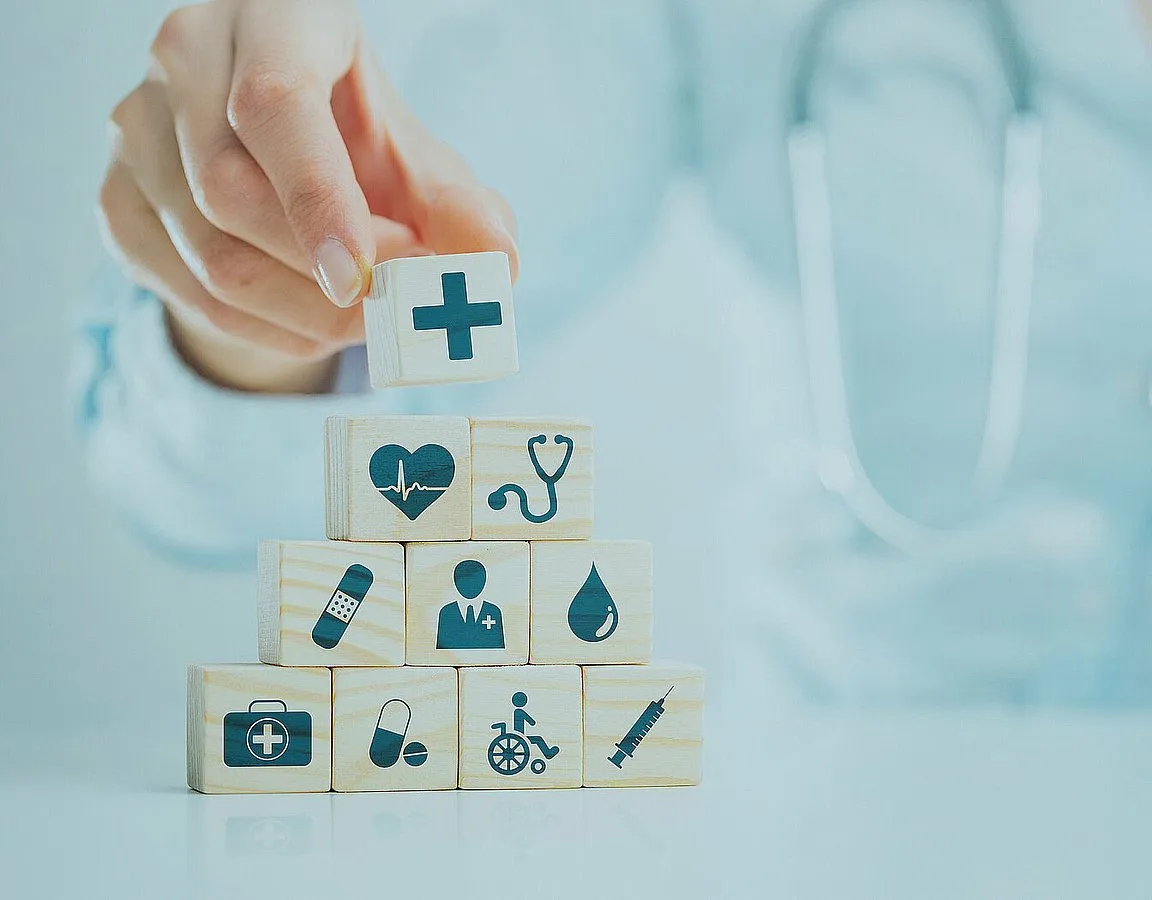 Eine Hand stapelt Holzklötze mit medizinischen Symbolen wie Kreuz, Herz und Stethoskop.
