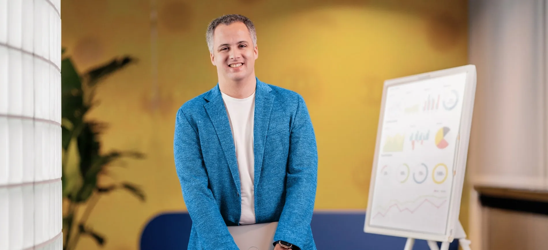 Ein lächelnder Mann im blauen Sakko hält ein Laptop vor einem Whiteboard mit IT-Diagrammen im modernen Büro.