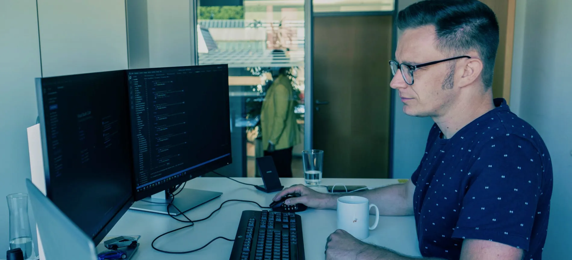 Du siehst einen Kollegen bei Peras GmbH, der konzentriert an zwei Monitoren im modernen Büro arbeitet.