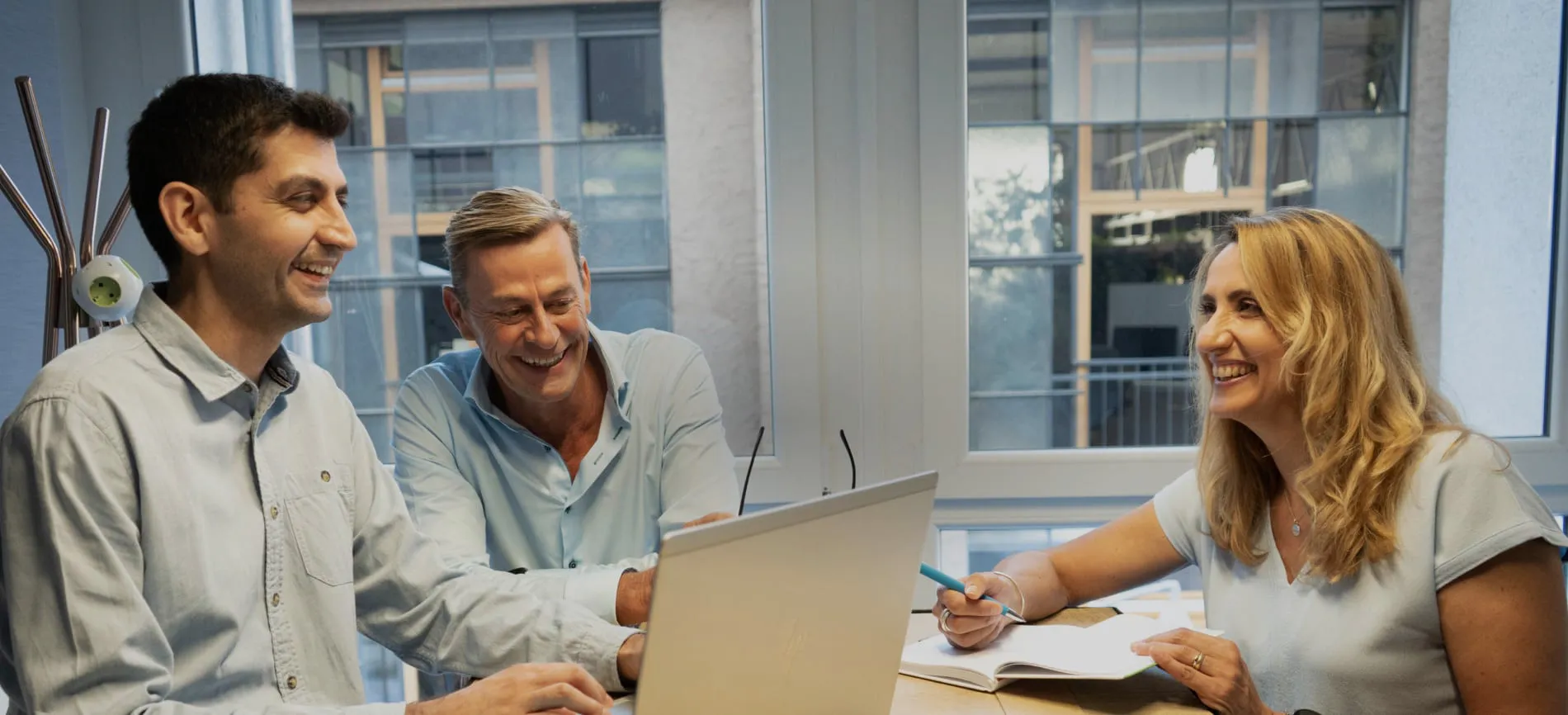 Drei Kolleg:innen lachen gemeinsam bei einer Besprechung im modernen Büro von Peras GmbH.
