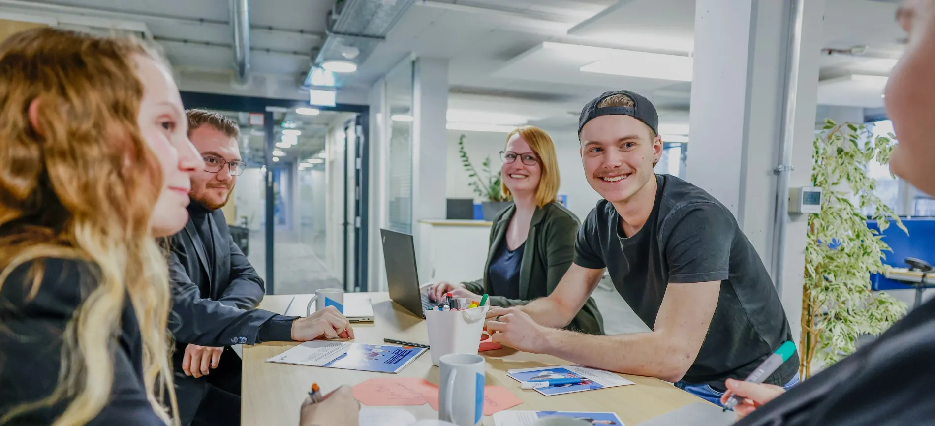 Mehrere junge Menschen sitzen gemeinsam am Tisch im modernen Büro und arbeiten im Team, alle wirken motiviert und freundlich.