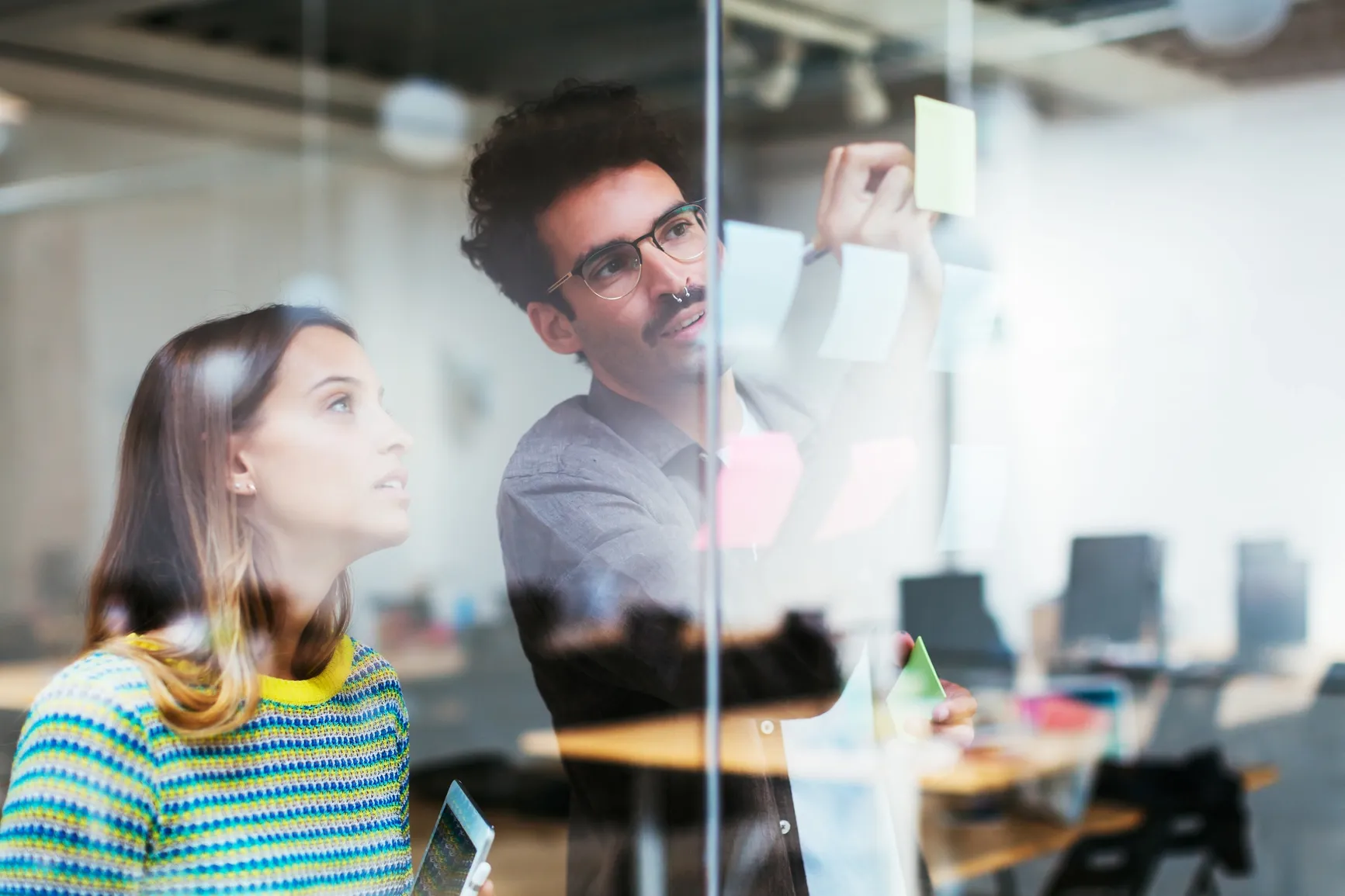 Two teammates brainstorm ideas with colorful sticky notes on a glass wall in a modern Ratepay office.