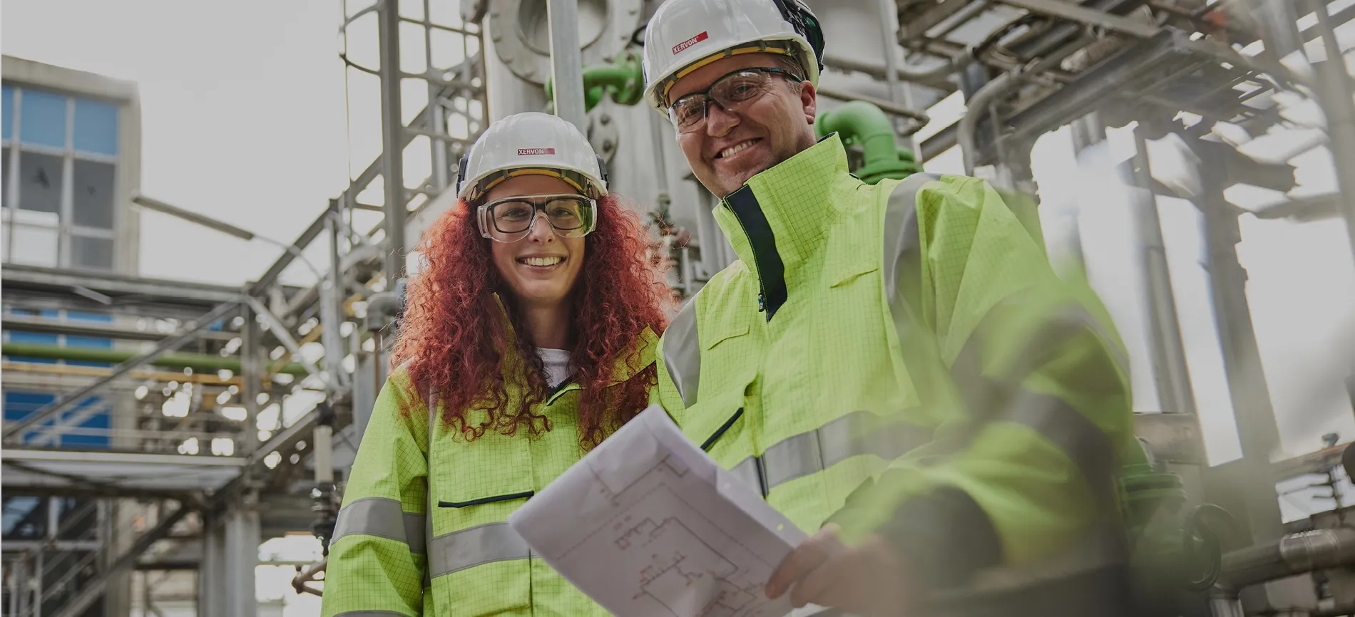 Zwei lächelnde Mitarbeitende in Schutzkleidung und Helmen stehen im Industrieanlagenbereich und halten einen Bauplan.