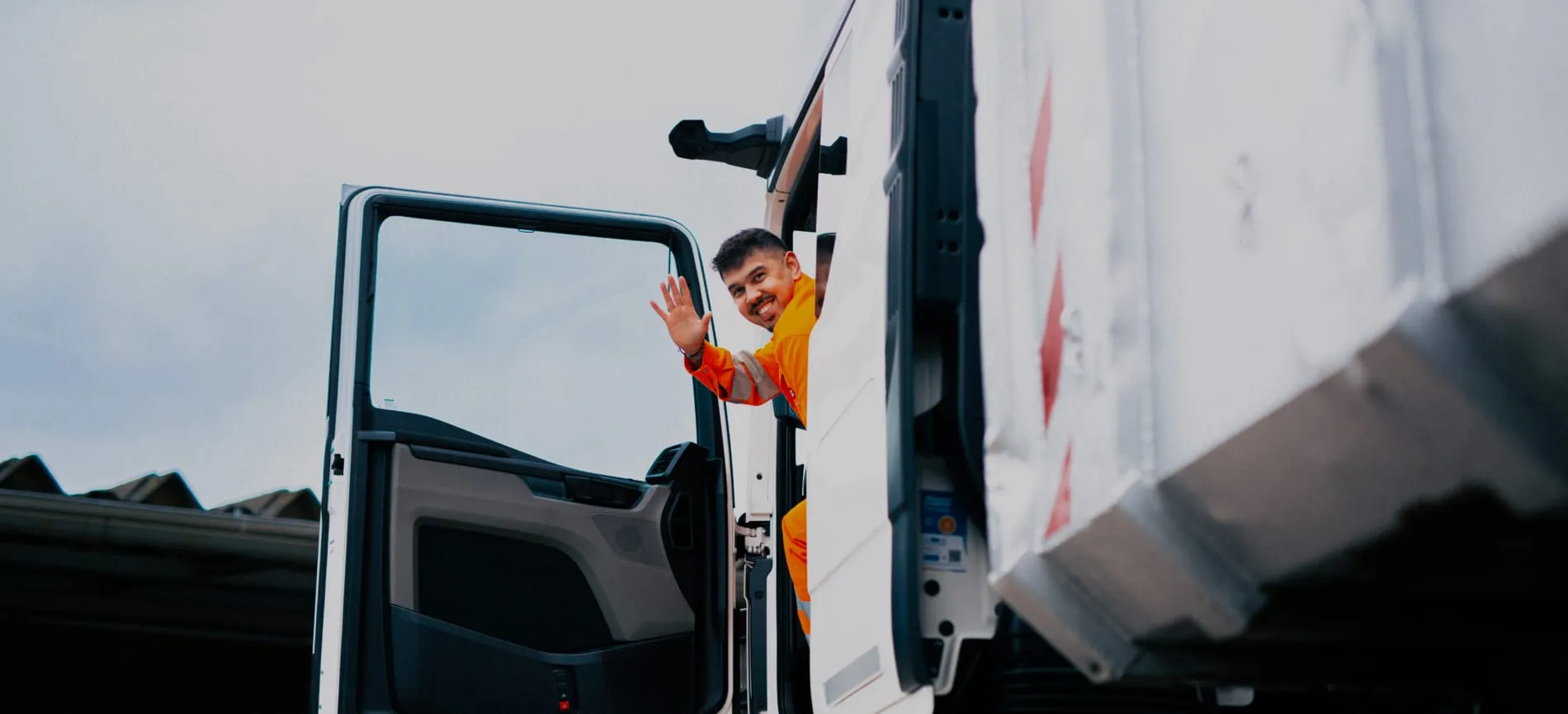 Ein Mitarbeiter in orangefarbener Arbeitskleidung winkt freundlich aus dem Fahrerhaus eines Lkw.