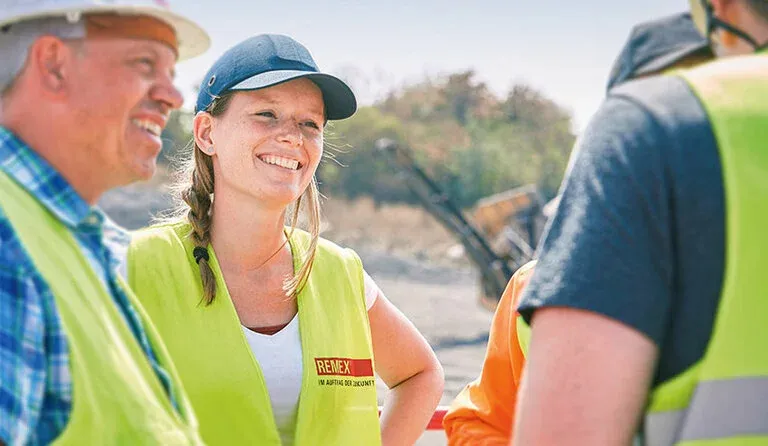 Junge Frau im REMEX-Sicherheitswesten lacht mit Kollegen auf einer Baustelle im Bereich Energie und Versorgungswirtschaft.