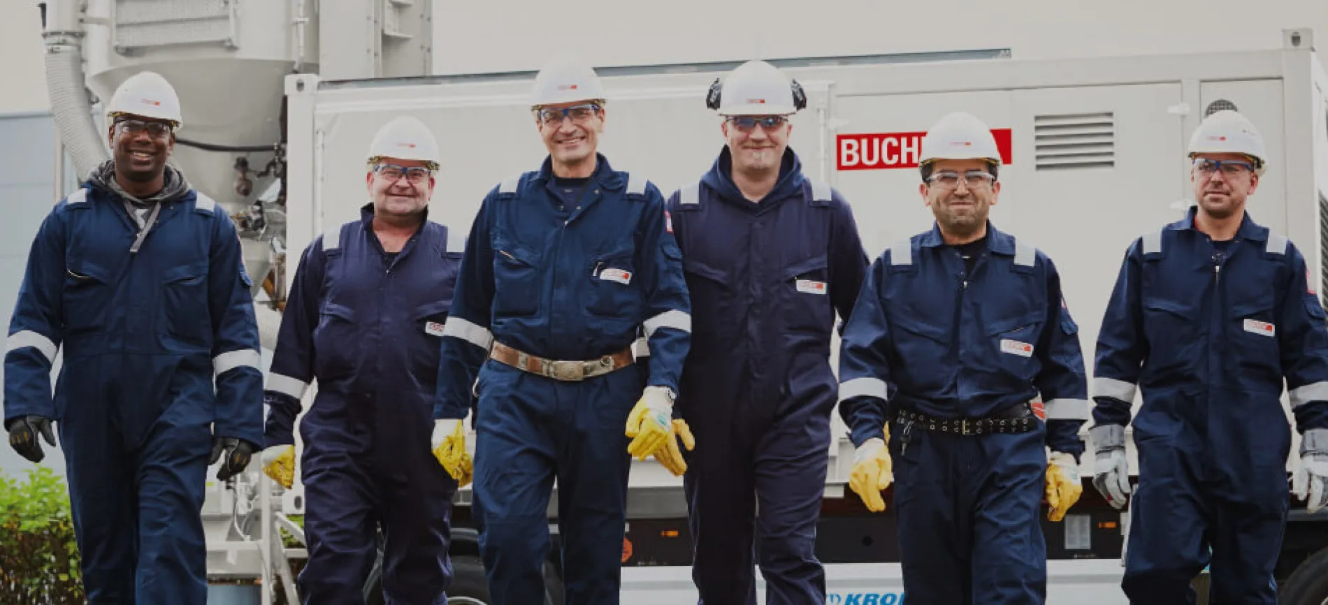 Zwei lächelnde Mitarbeitende in Schutzkleidung und Helmen stehen im Industrieanlagenbereich und halten einen Bauplan.