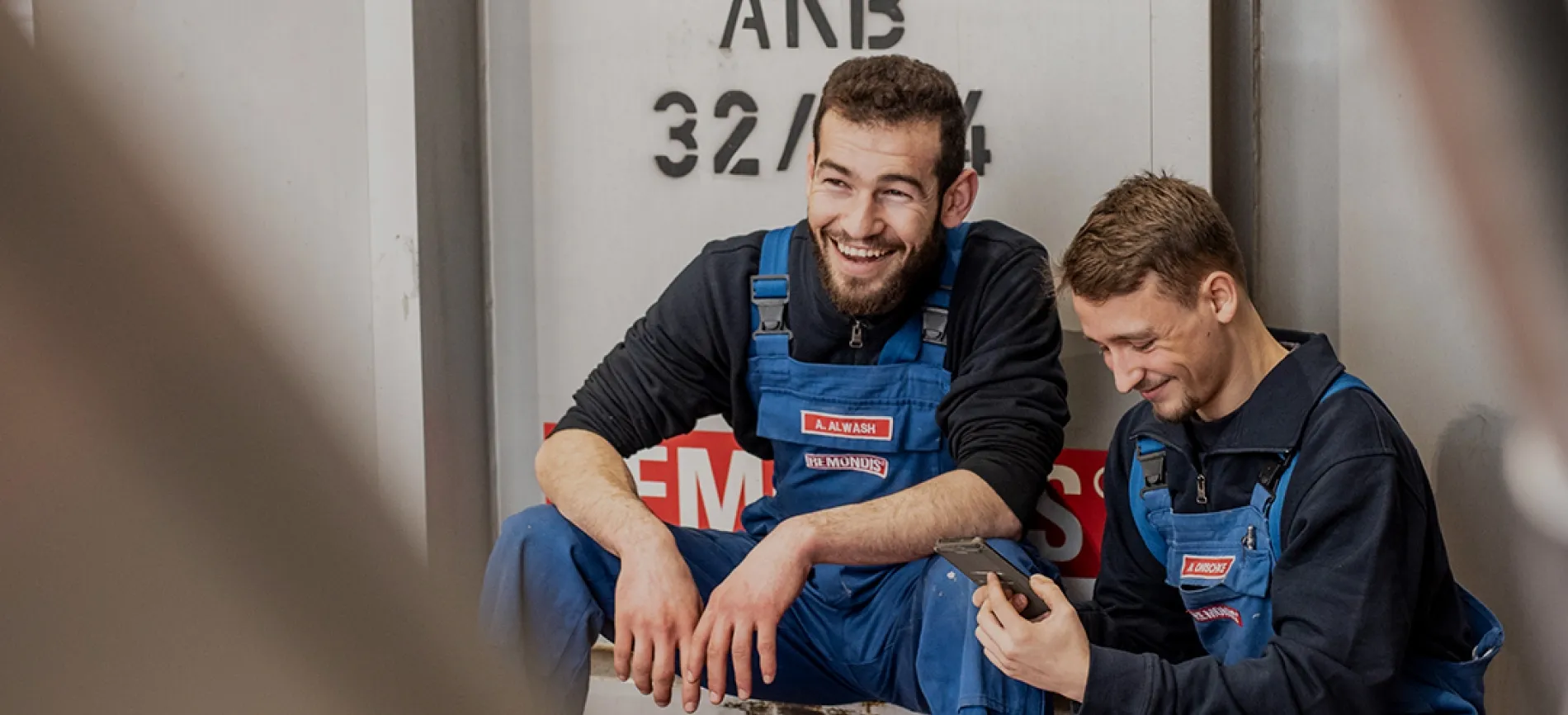Zwei junge REMONDIS-Mitarbeiter in blauer Arbeitskleidung lachen gemeinsam in einer Pause und schauen auf ein Handy.