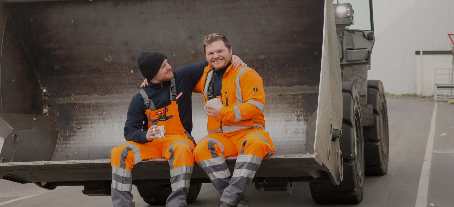 Zwei Kollegen in orangefarbener Arbeitskleidung machen gemeinsam Pause auf einer Bagger-Schaufel und lachen.