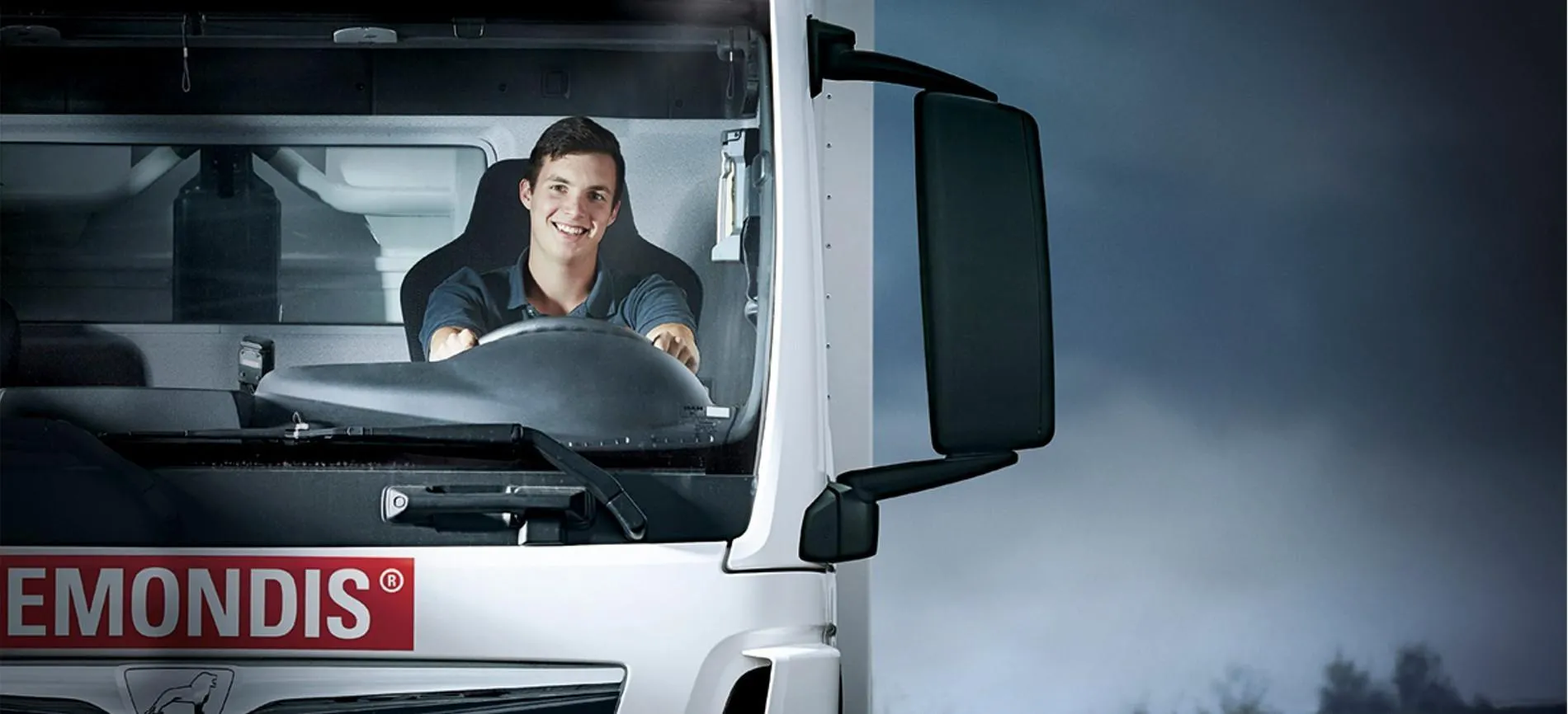 Ein junger Mann sitzt lächelnd am Steuer eines REMONDIS-Lkw und blickt direkt in die Kamera.