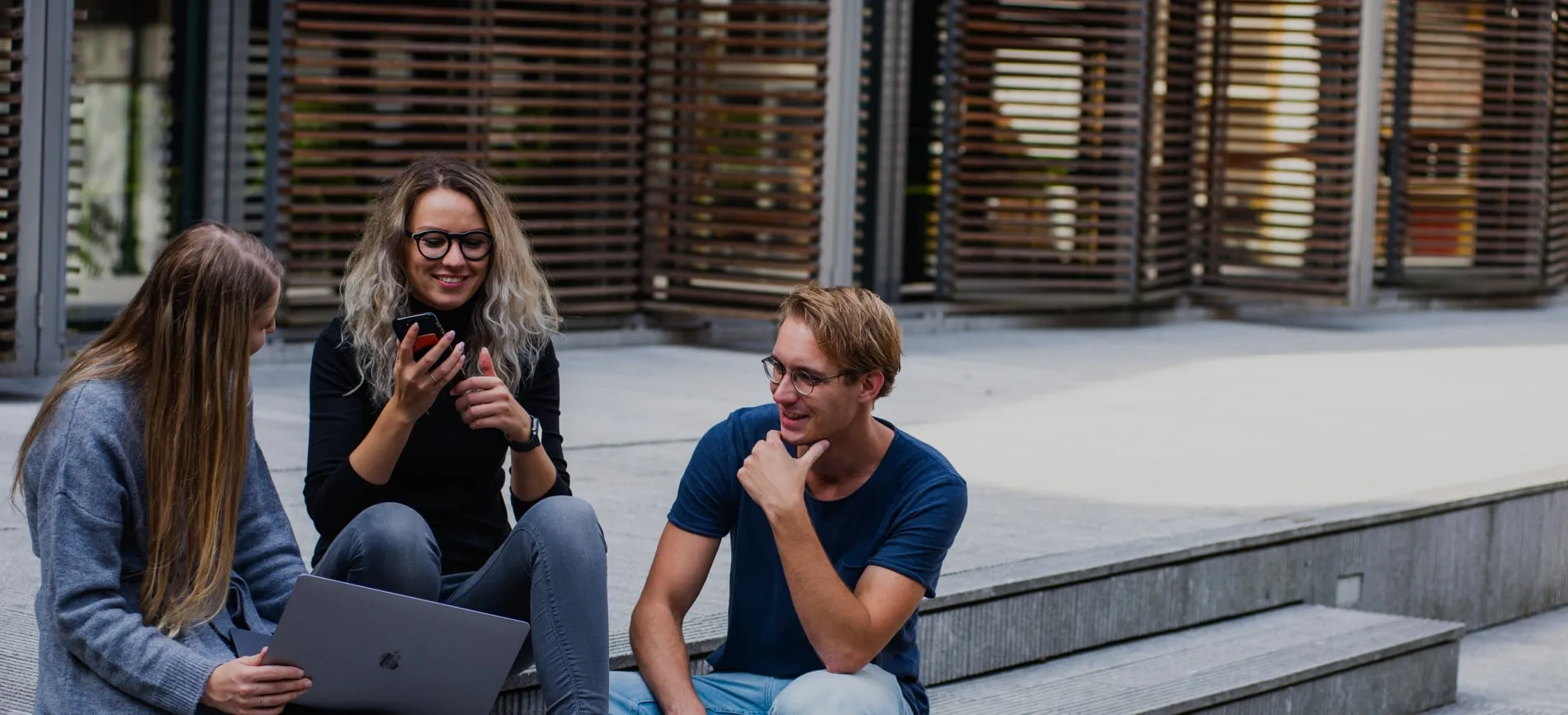 Drei junge Menschen sitzen draußen, lachen und arbeiten gemeinsam mit Laptop und Smartphone an einem Projekt.