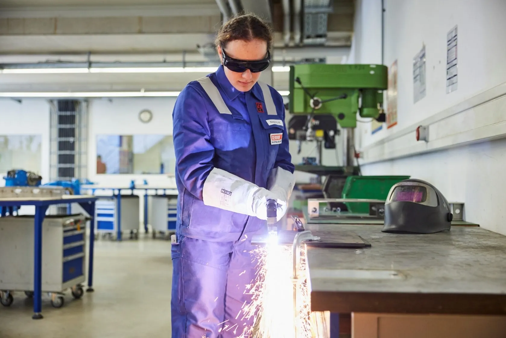 Eine Auszubildende bearbeitet mit Schutzbrille und Schutzkleidung ein Metallstück, während Funken entstehen.