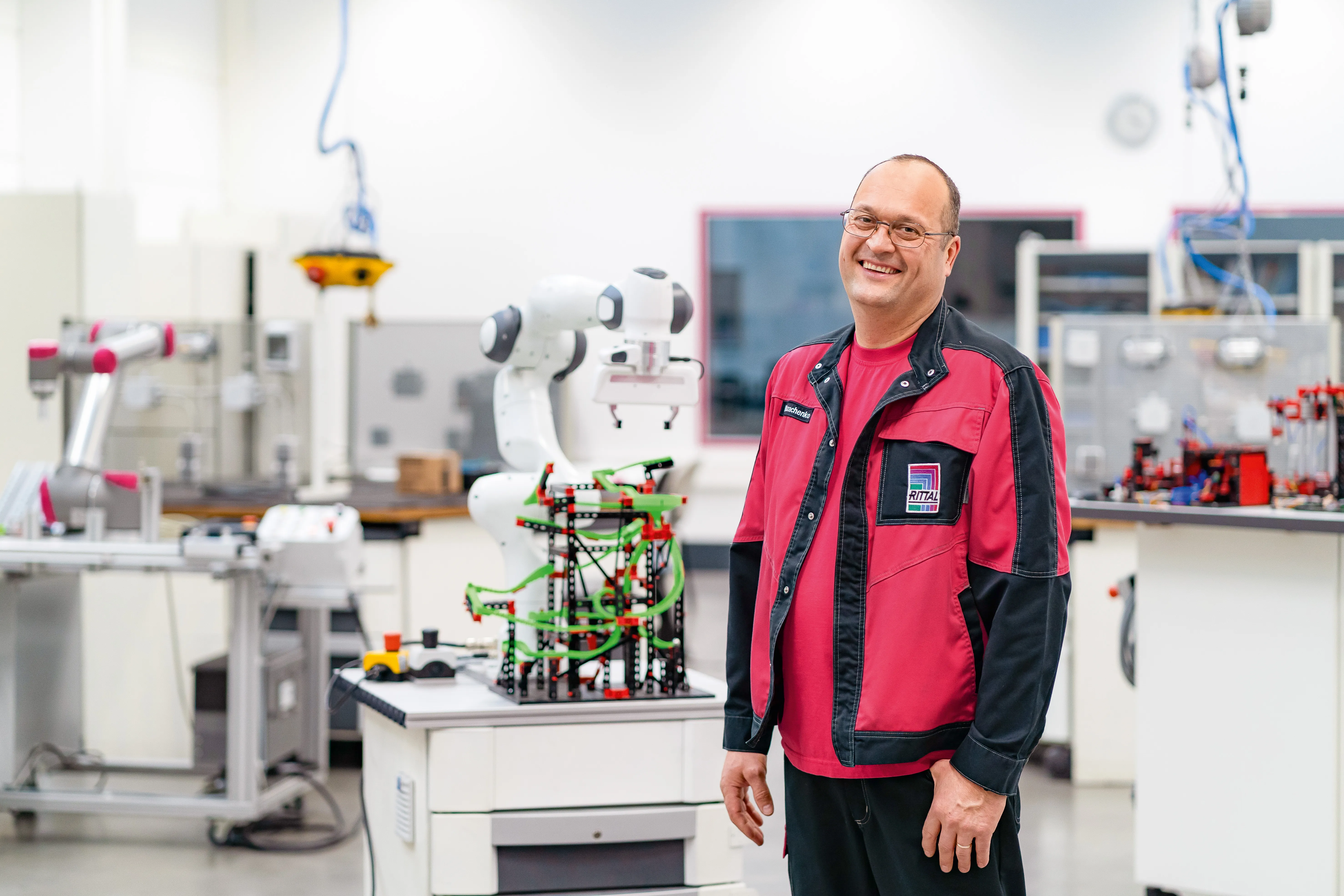 A friendly Rittal employee in a vibrant lab, smiling next to high-tech robotics equipment.