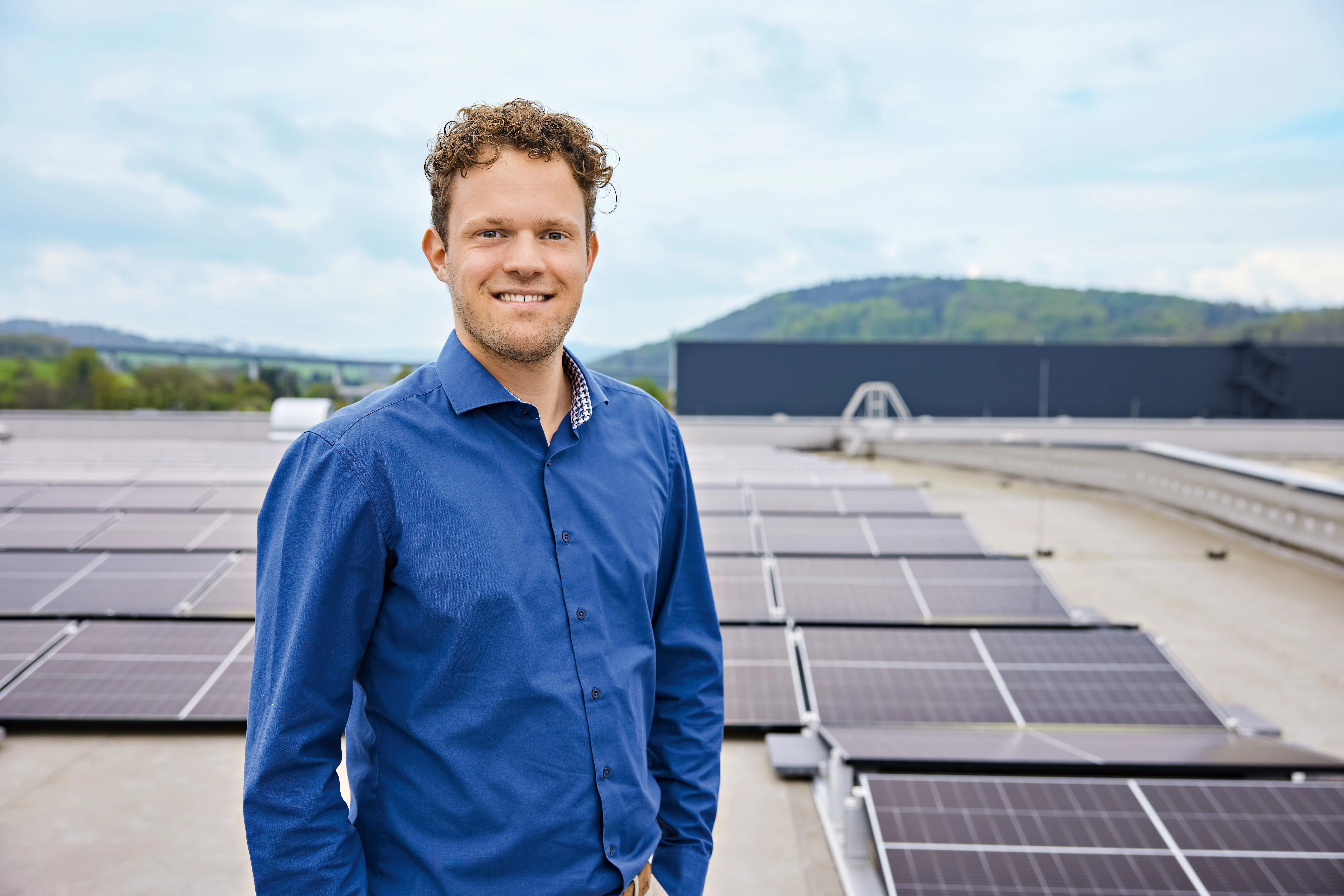 Ein Mann in blauem Hemd steht auf einem Dach mit Solarpanelen im Hintergrund und lächelt in die Kamera.