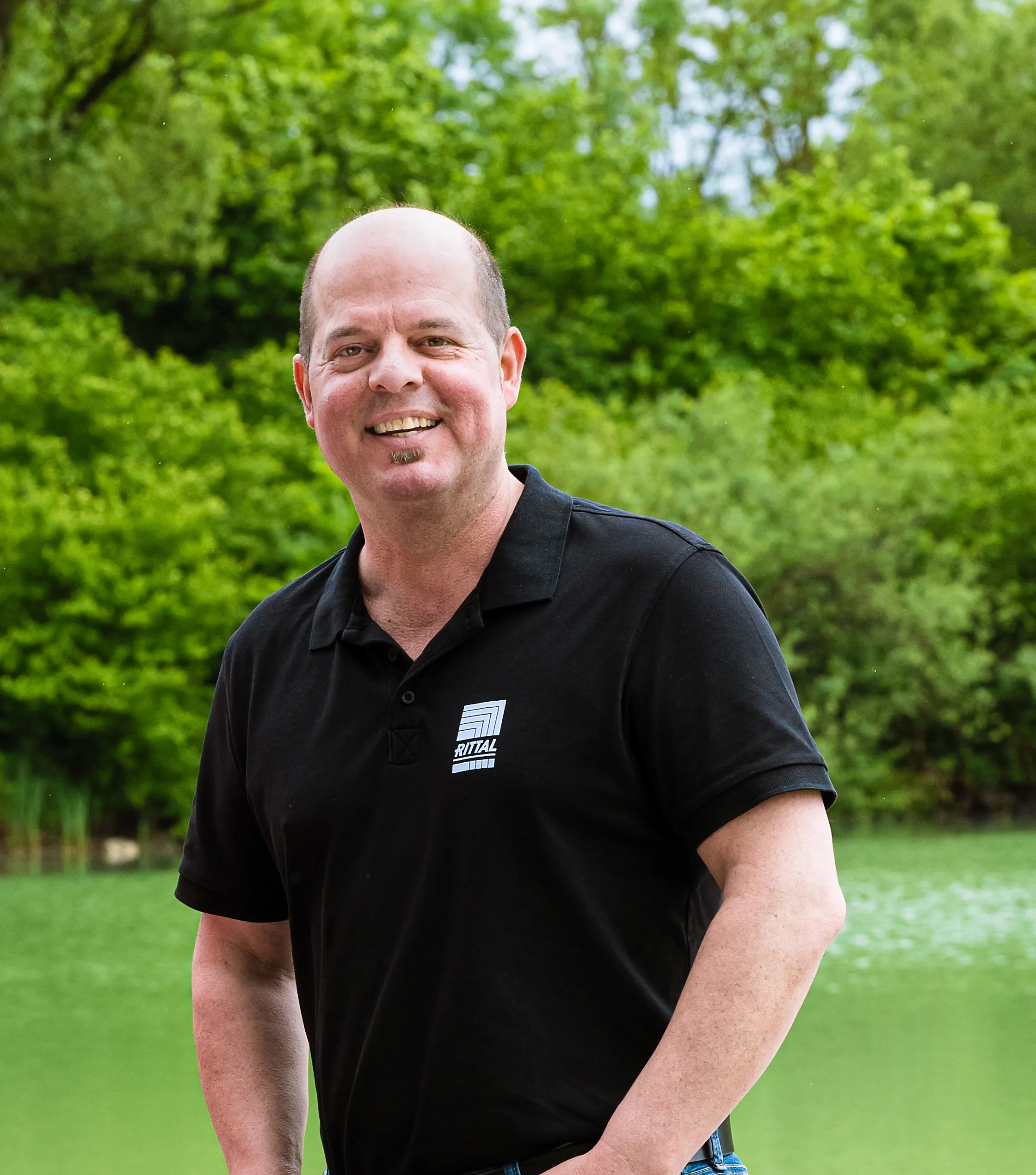 Ein lächelnder Mann in einem Rittal-Polohemd steht vor einer grünen Landschaft.