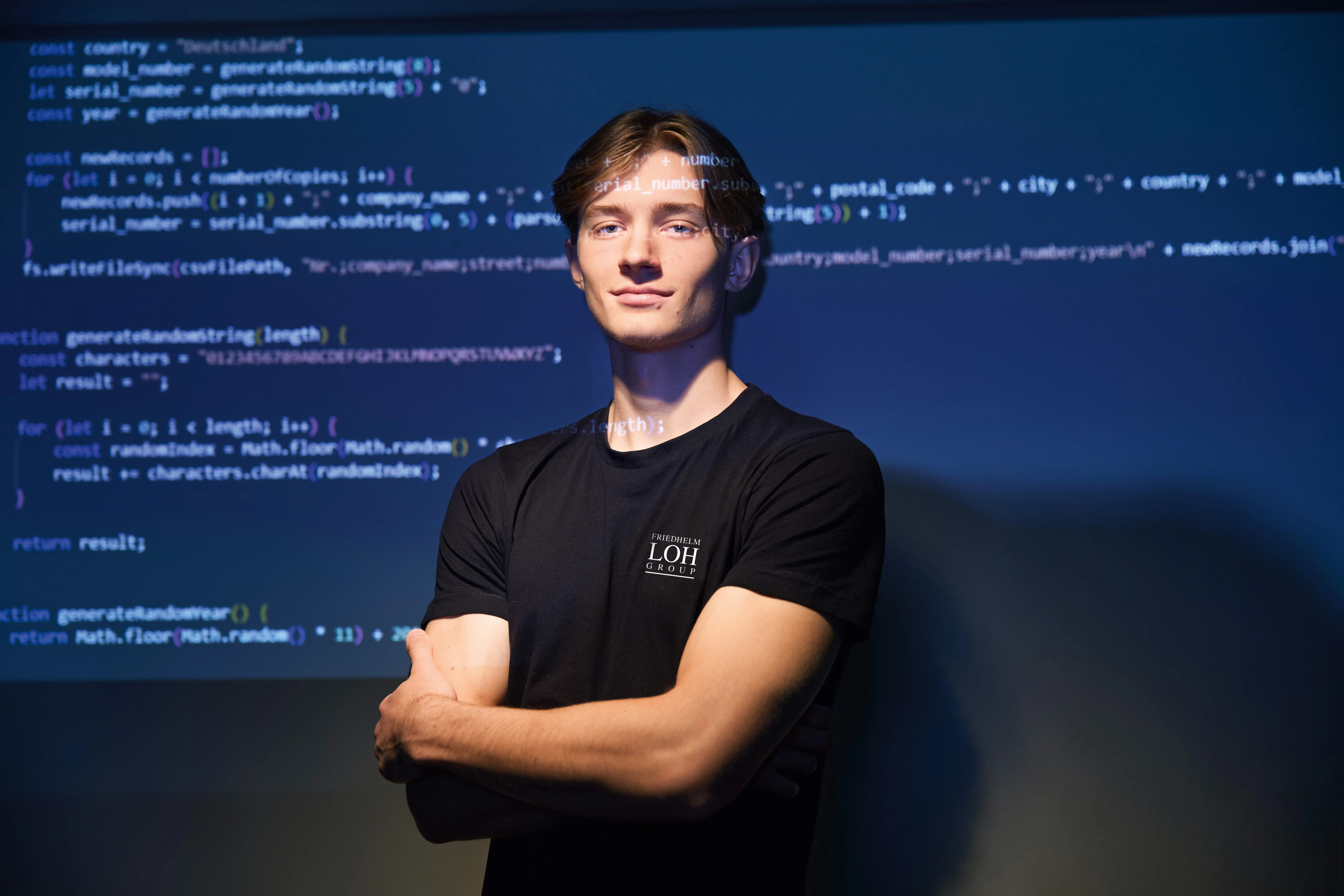 Young software developer confidently standing with arms crossed, code projected on screen behind, showcasing tech skills at Rittal GmbH & Co. KG.