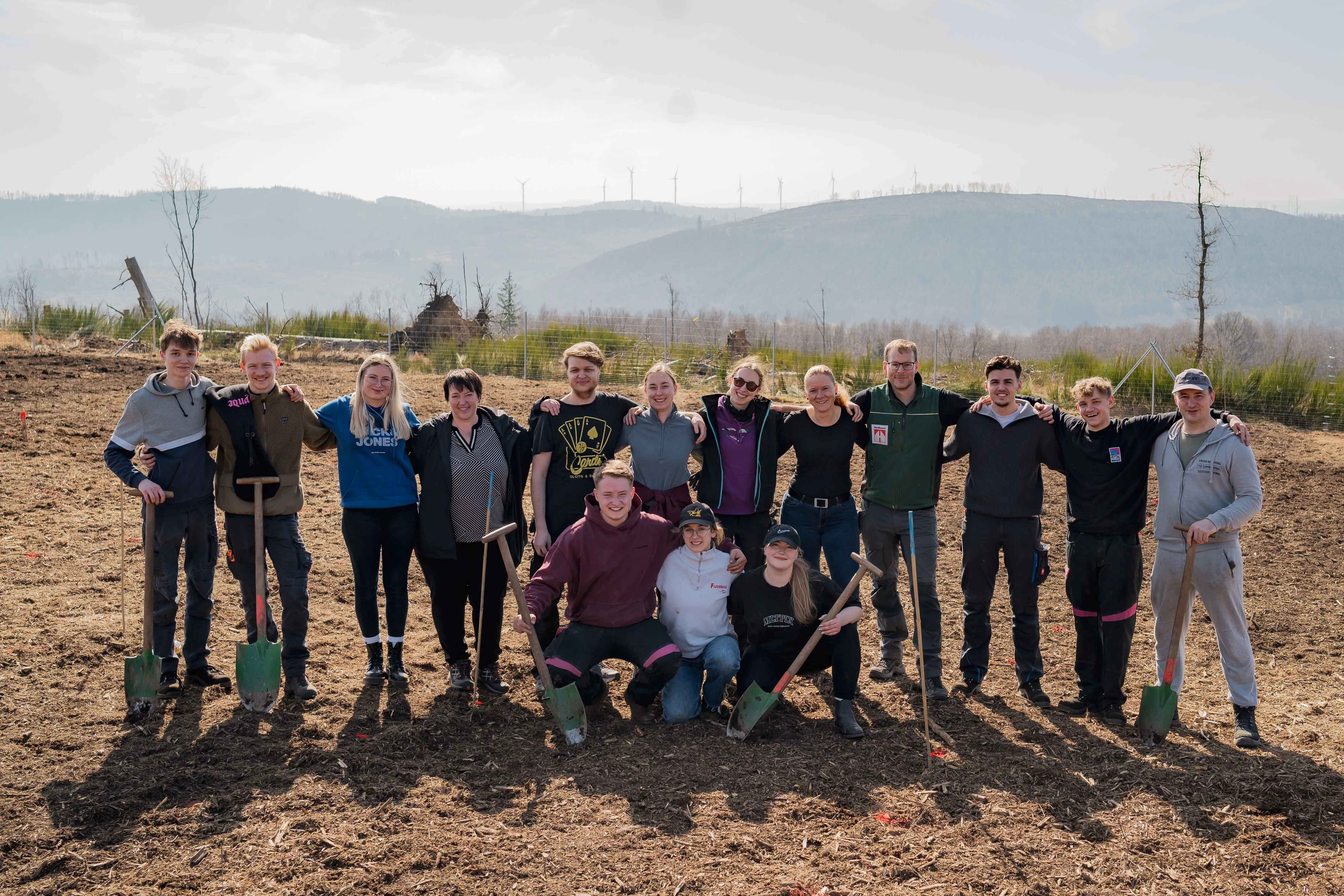 Team Rittal enjoying a day outdoors, planting trees and bonding under a sunny sky, with wind turbines in the distance.
