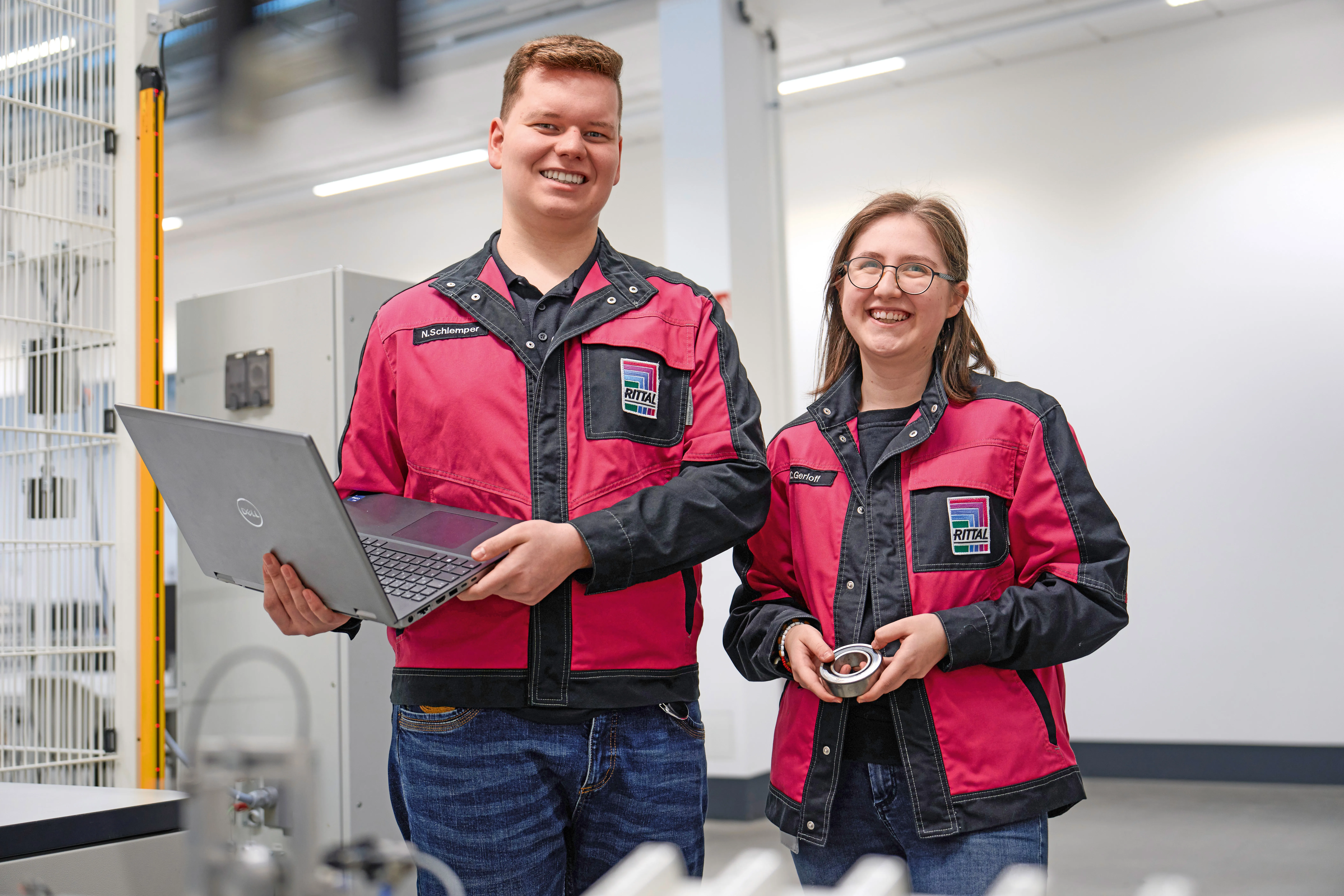 Two Rittal employees in red jackets smiling, one holding a laptop, in a tech workspace.