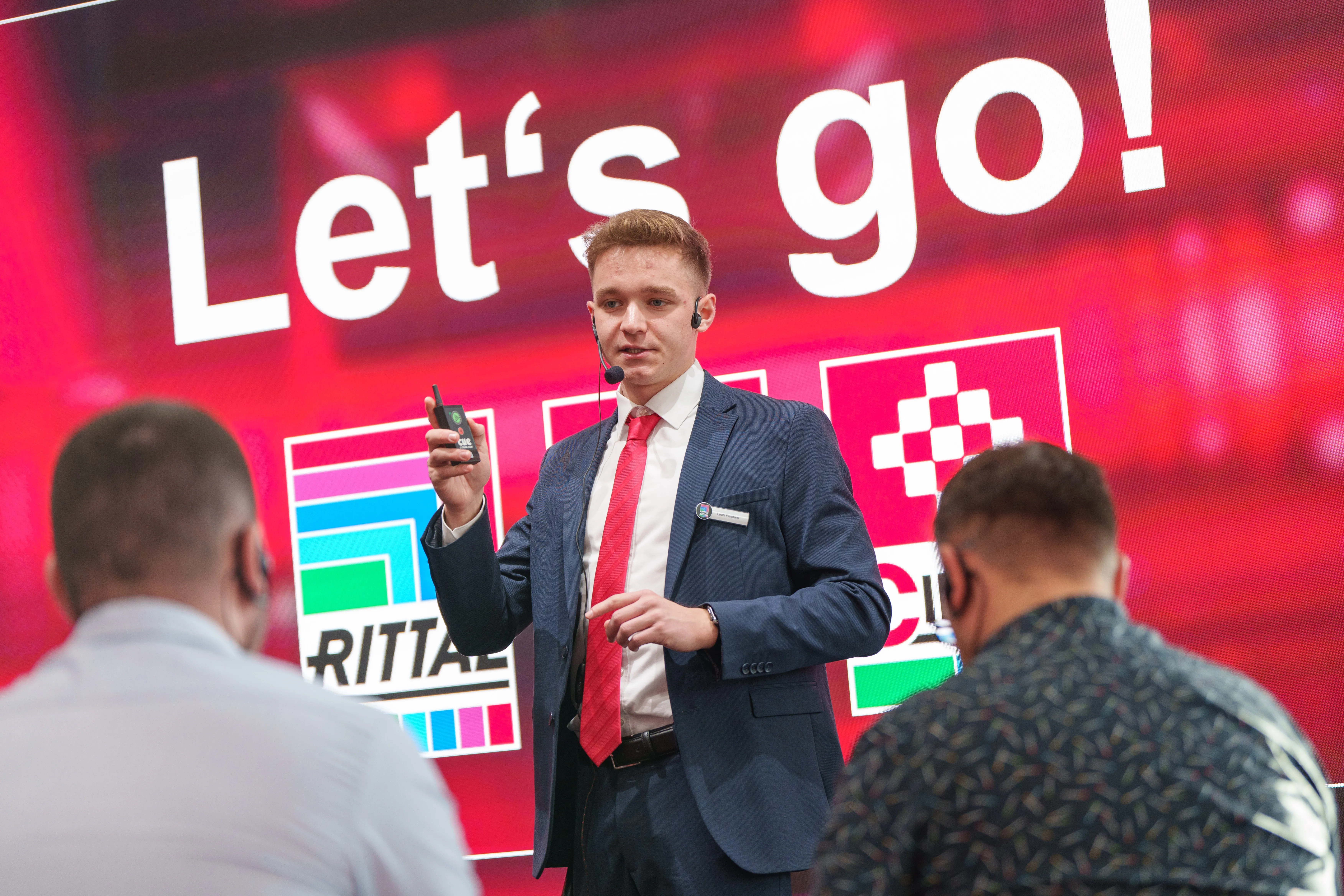 A confident speaker in a suit engages an audience at a Rittal IT event, with "Let's go!" displayed on a vibrant screen.