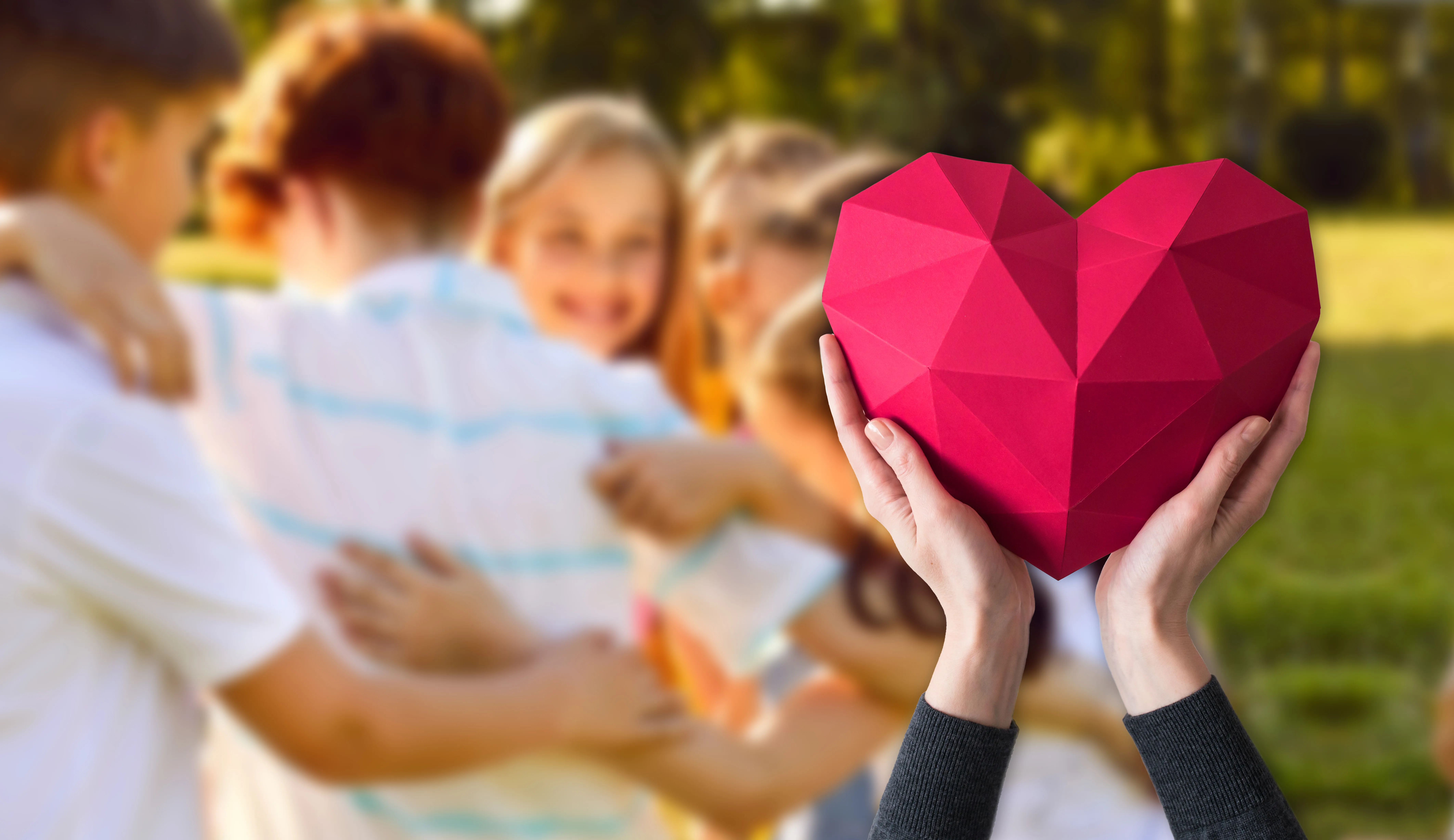 Hände halten ein rotes, geometrisches Herz, im Hintergrund umarmen sich Menschen. Symbol für Teamarbeit und Zusammenhalt.