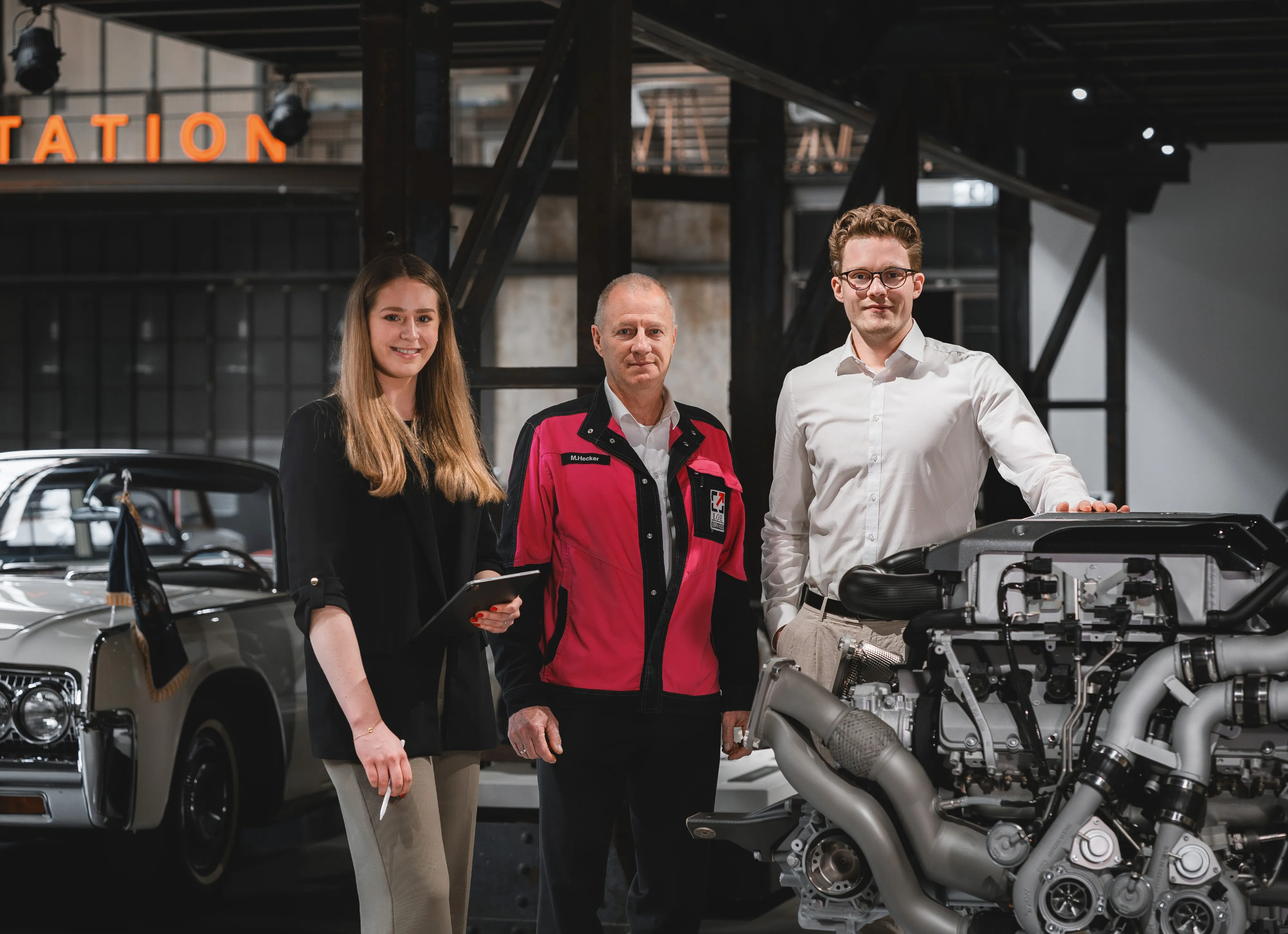Drei Personen stehen in einer industriellen Umgebung, neben einem Motor und einem klassischen Auto, und lächeln in die Kamera.