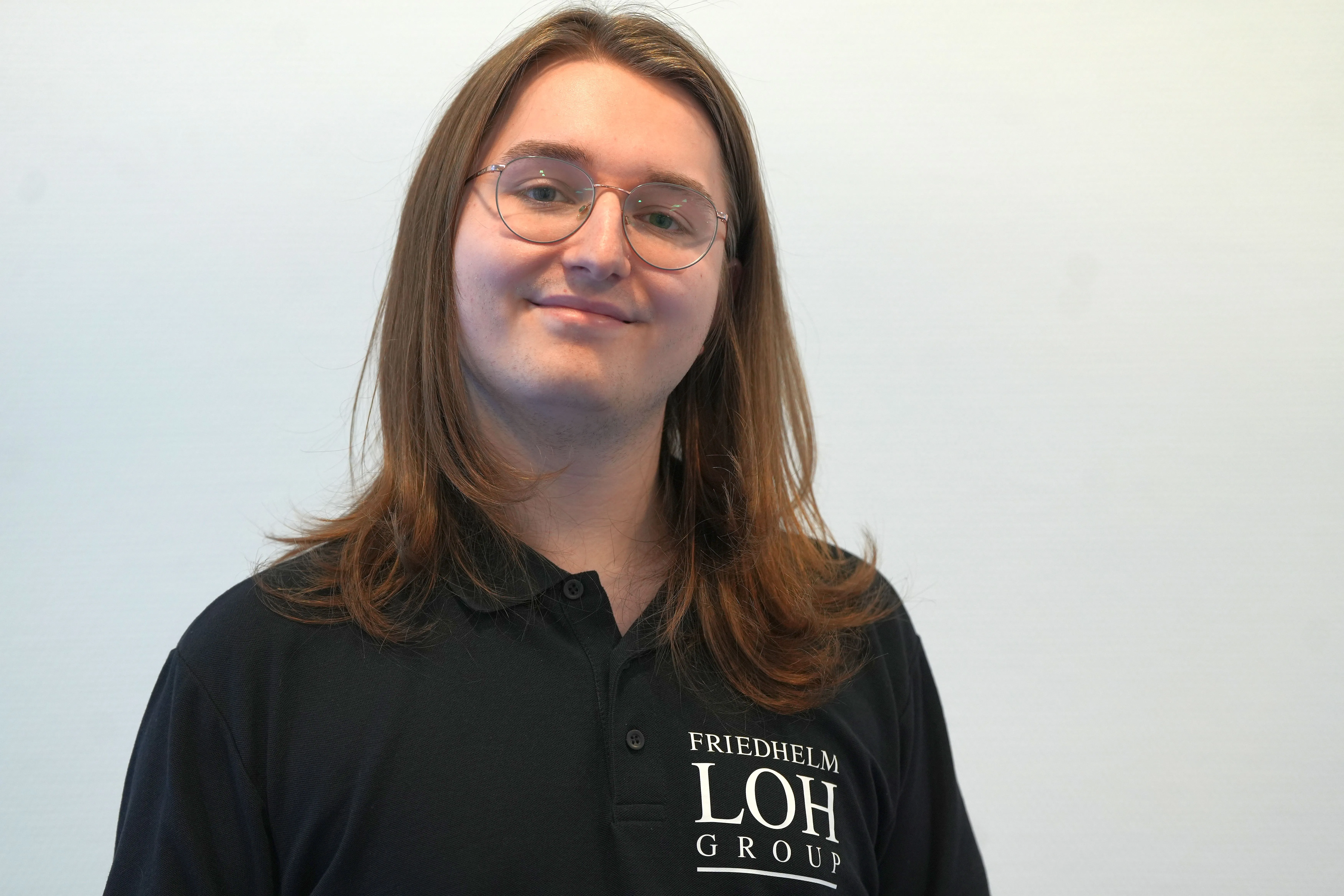 A young man in sunglasses smiles while taking a selfie on a sunny beach, with trees and a clear sky in the background.