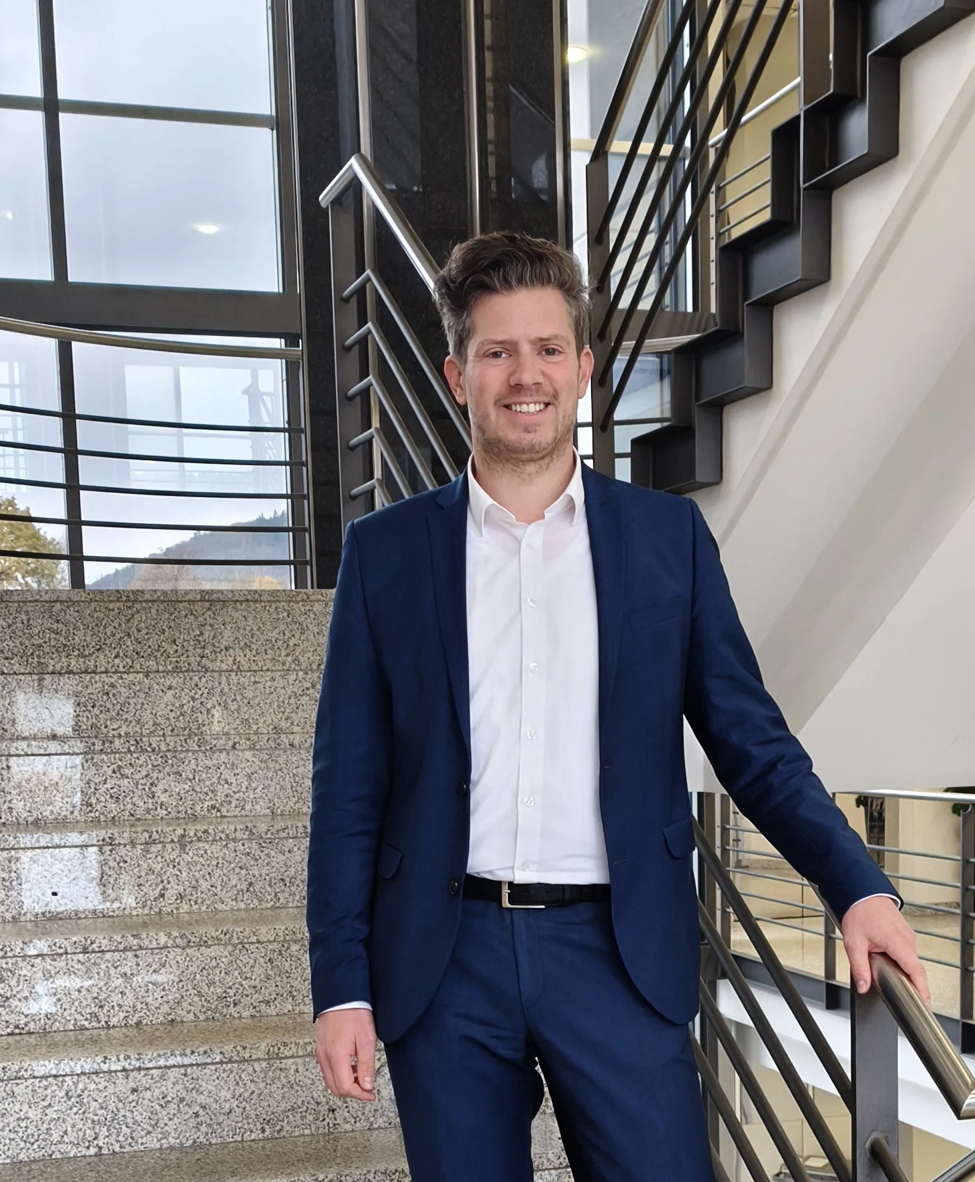 Smiling man in a blue suit standing on a modern staircase at Rittal GmbH & Co. KG, showcasing a professional IT environment.