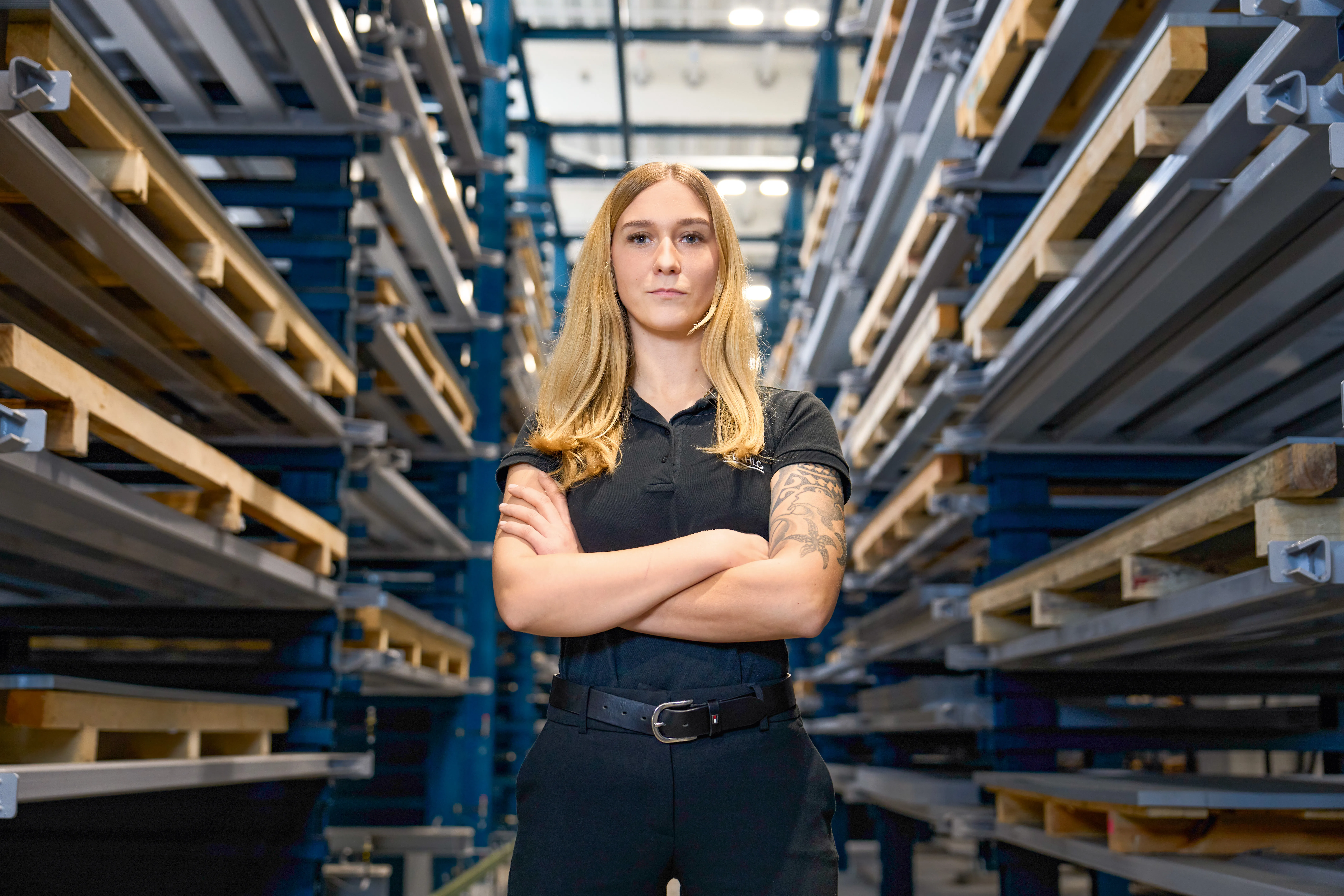 Eine Frau in schwarzer Arbeitskleidung steht mit verschränkten Armen in einem Lager bei Rittal GmbH & Co. KG.