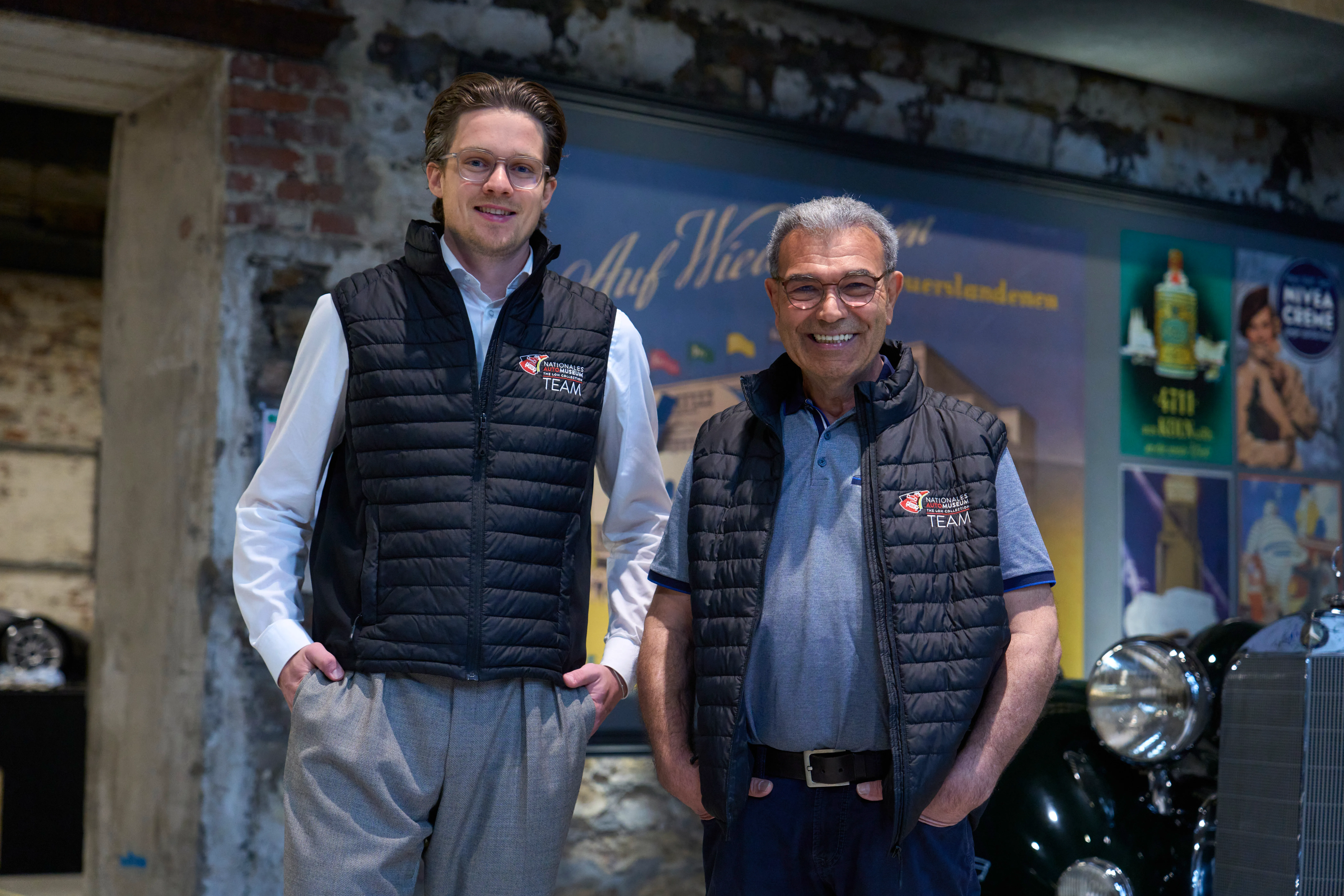 Zwei Männer in Teamwesten lächeln in die Kamera. Sie stehen vor einer Wand mit Werbeplakaten.