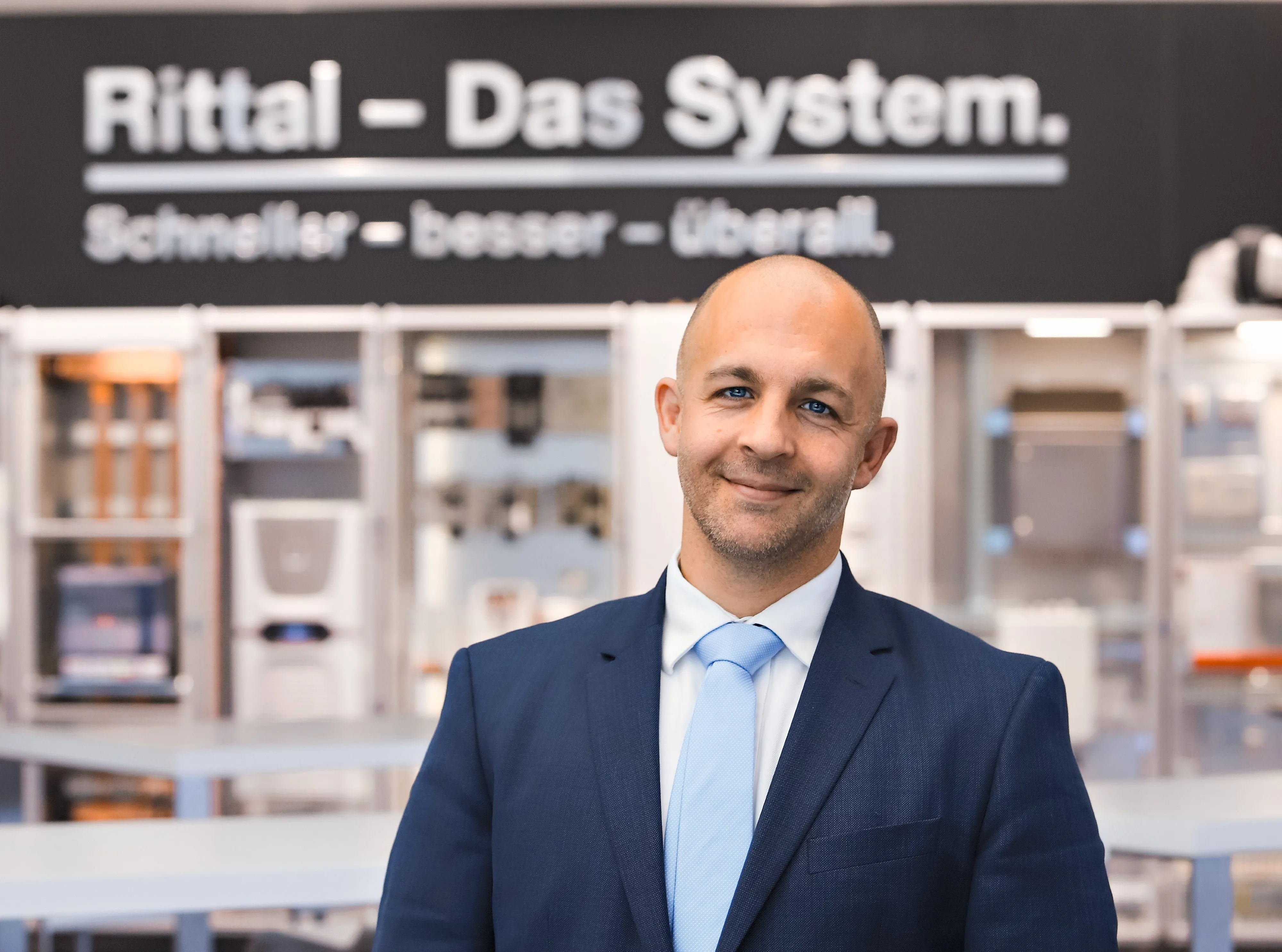Ein lächelnder Mann im Anzug steht vor einem Rittal-Schriftzug. Perfekt für Deine Karriere bei Rittal im IT-Bereich.