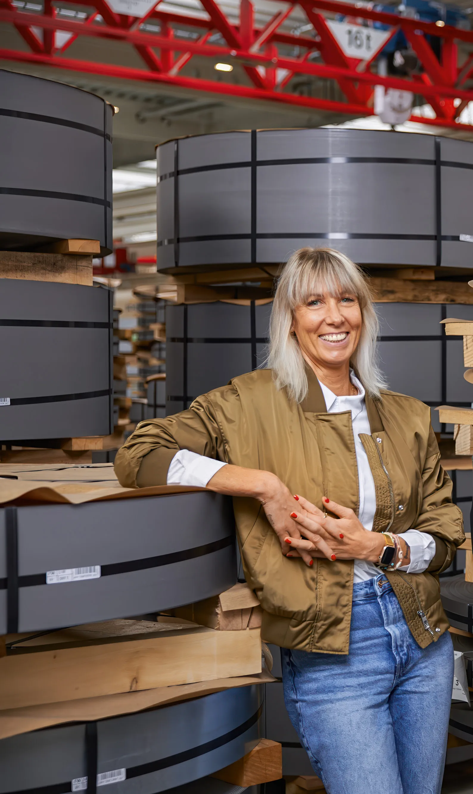 Eine Frau in einer Fabrikumgebung lehnt lächelnd an gestapelten Metallrollen. Sie trägt eine braune Jacke und Jeans.