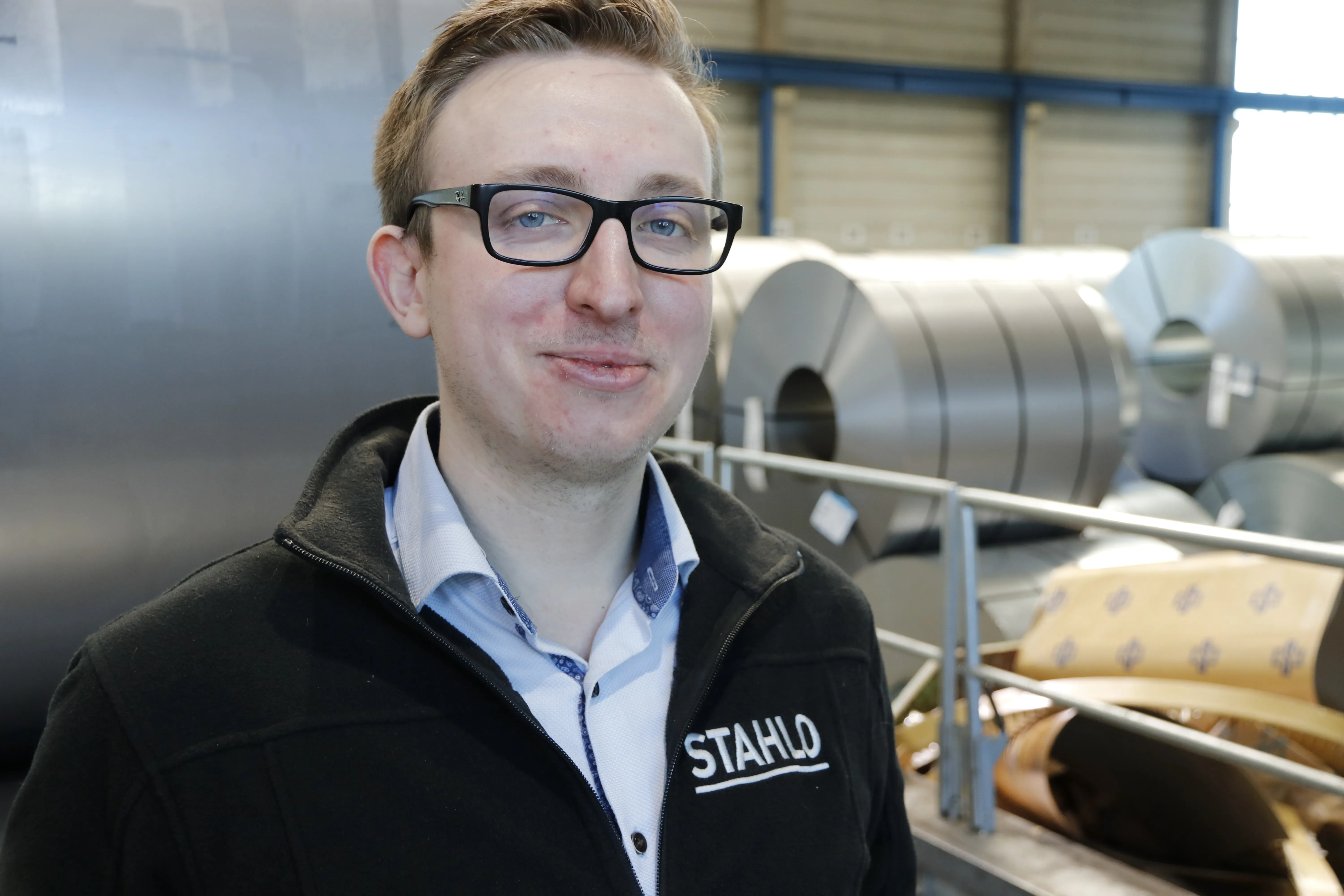 Ein Mann mit Brille lächelt in einer Produktionshalle. Er trägt eine Jacke mit dem Aufdruck "STAHLD". Im Hintergrund sind Metallrollen zu sehen.