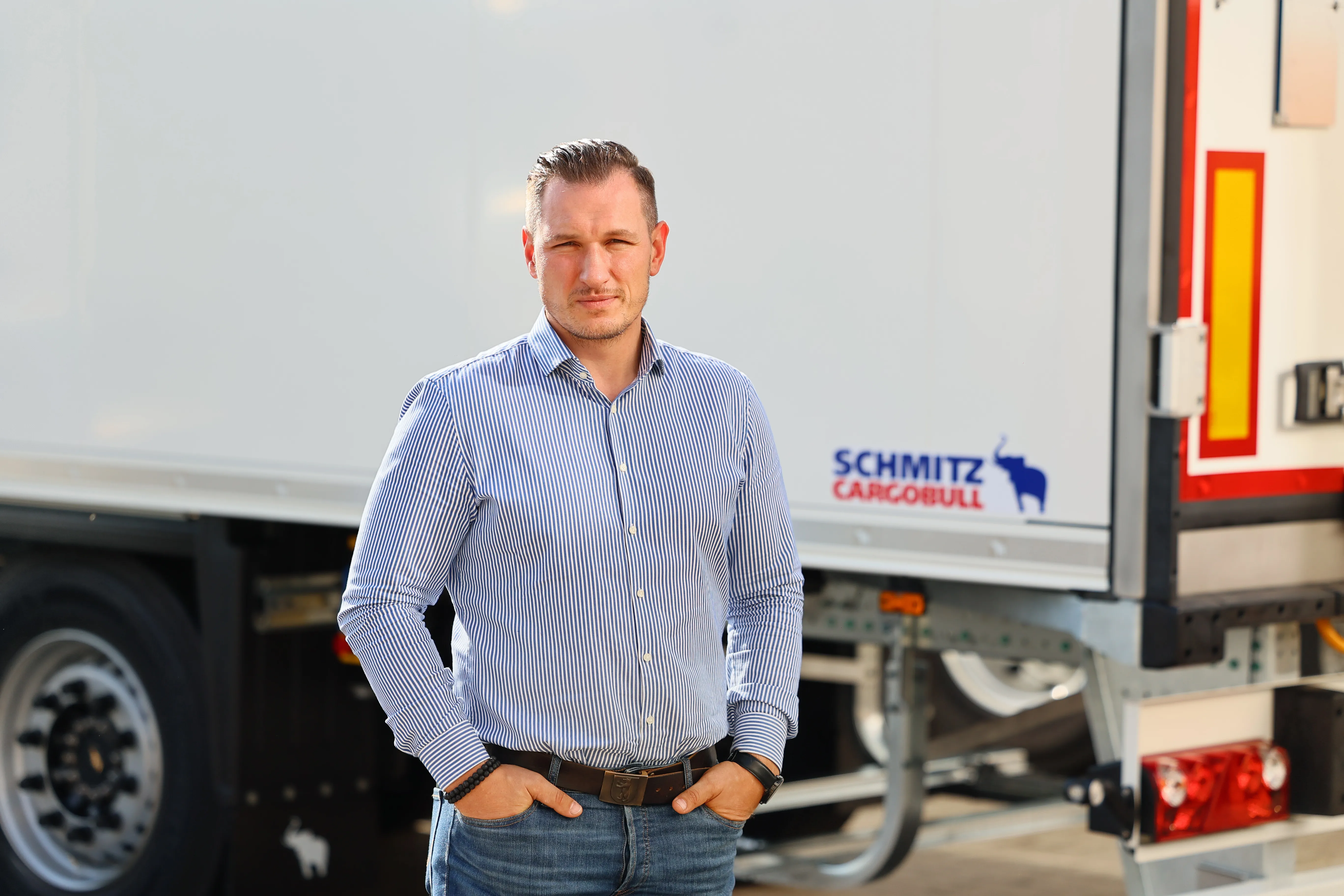 Confident person in casual shirt standing in front of a Schmitz Cargobull trailer, ready to take on the automotive world.