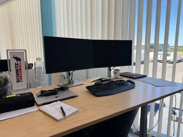 A stylish workstation at Schmitz Cargobull AG with dual monitors, notebook, and a branded sign, overlooking the outdoors.