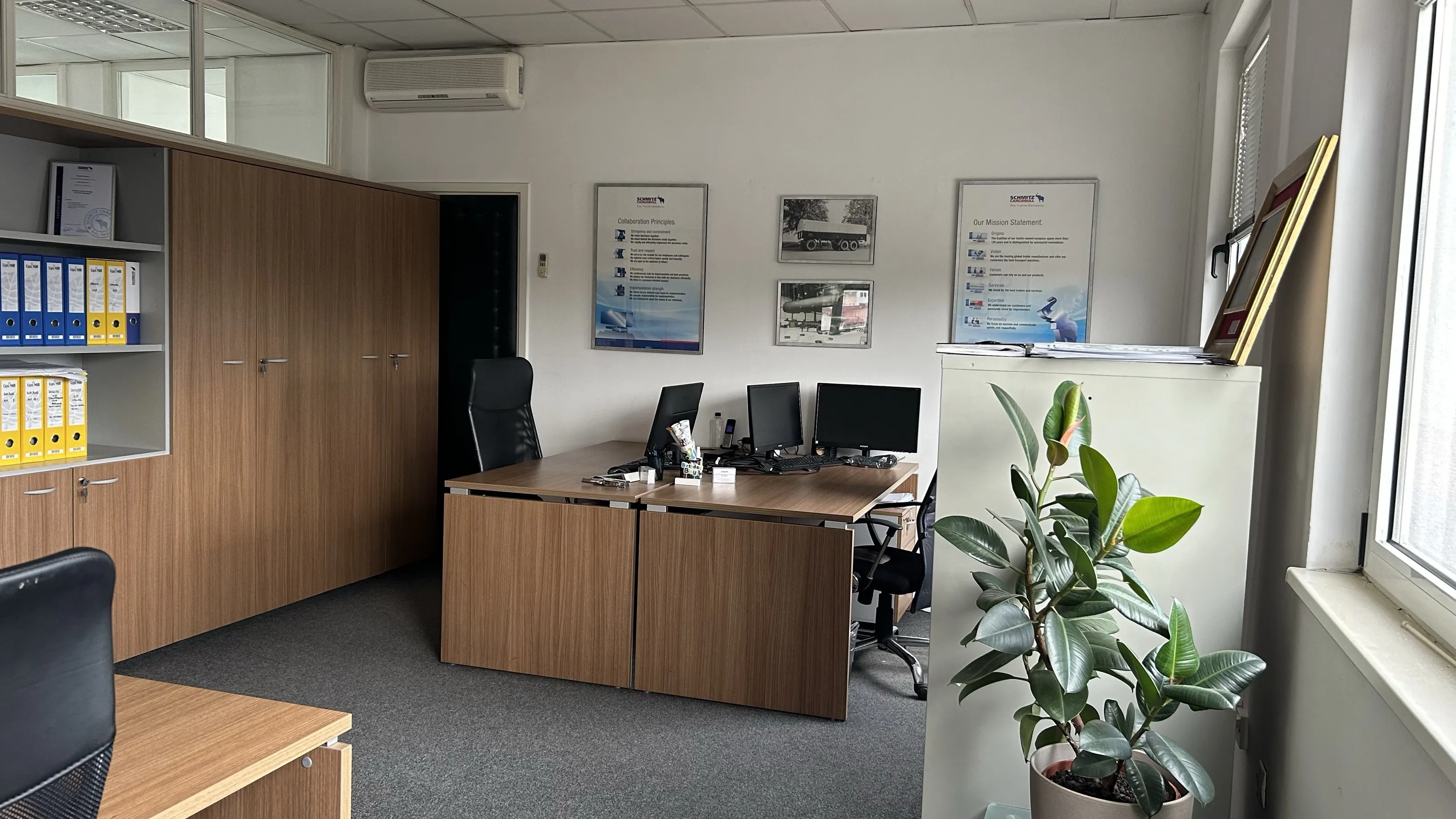 A cozy Schmitz Cargobull AG office with wooden desks, computers, cabinets filled with files, and a lively plant by the window.