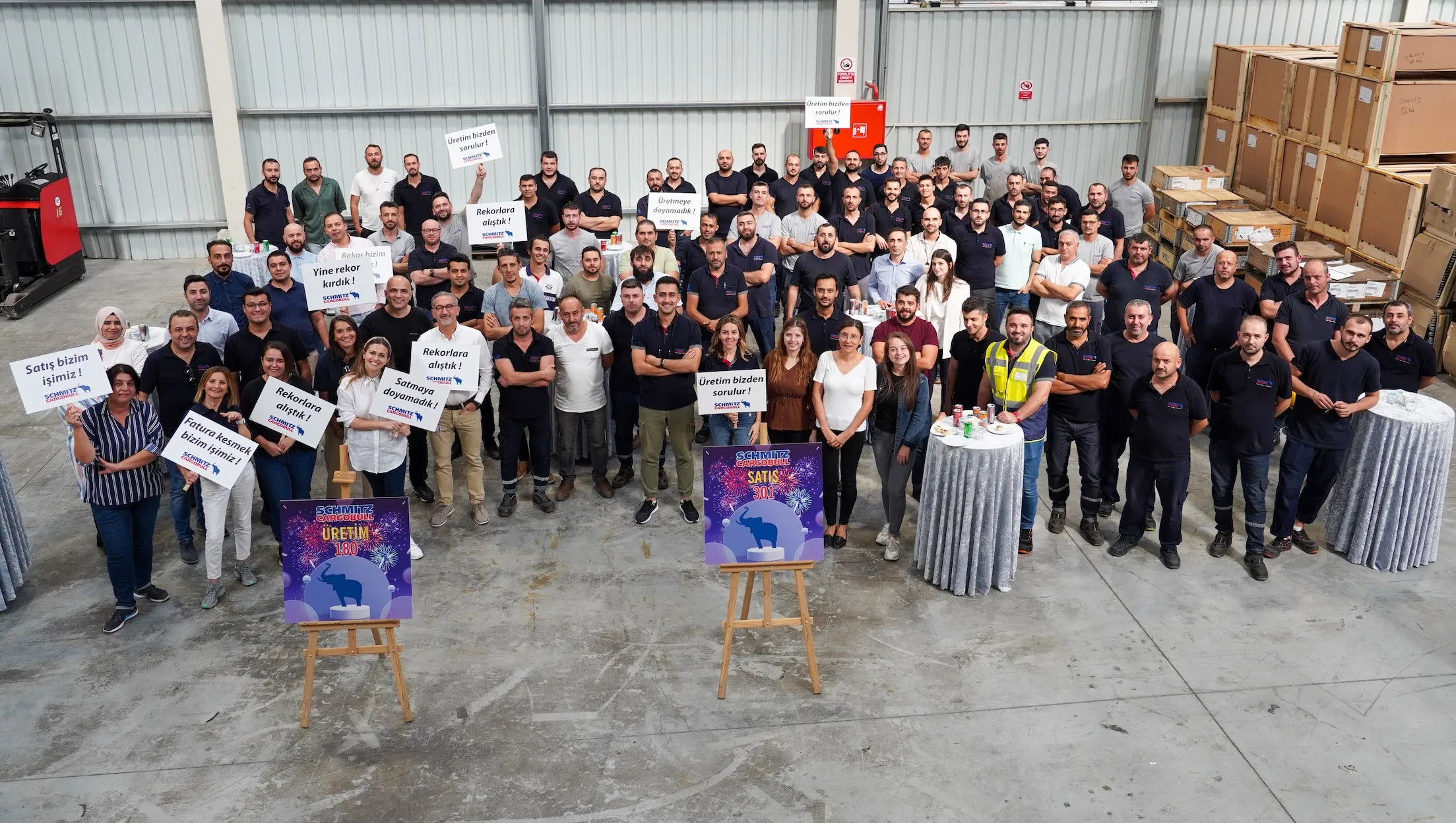 Schmitz Cargobull AG team celebrating a sales milestone with smiles and signs in a warehouse setting.