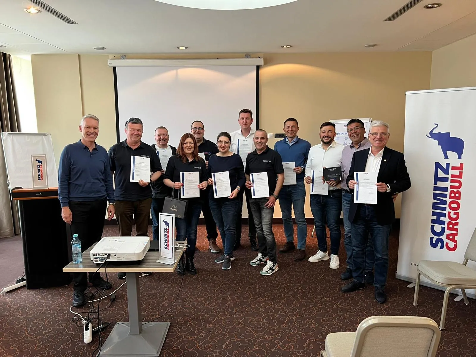 Schmitz Cargobull team celebrating with certificates, smiling and standing proudly in a conference room.