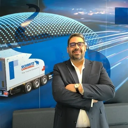 A smiling man in a suit stands in front of a Schmitz Cargobull truck graphic, ready to welcome you to the team.