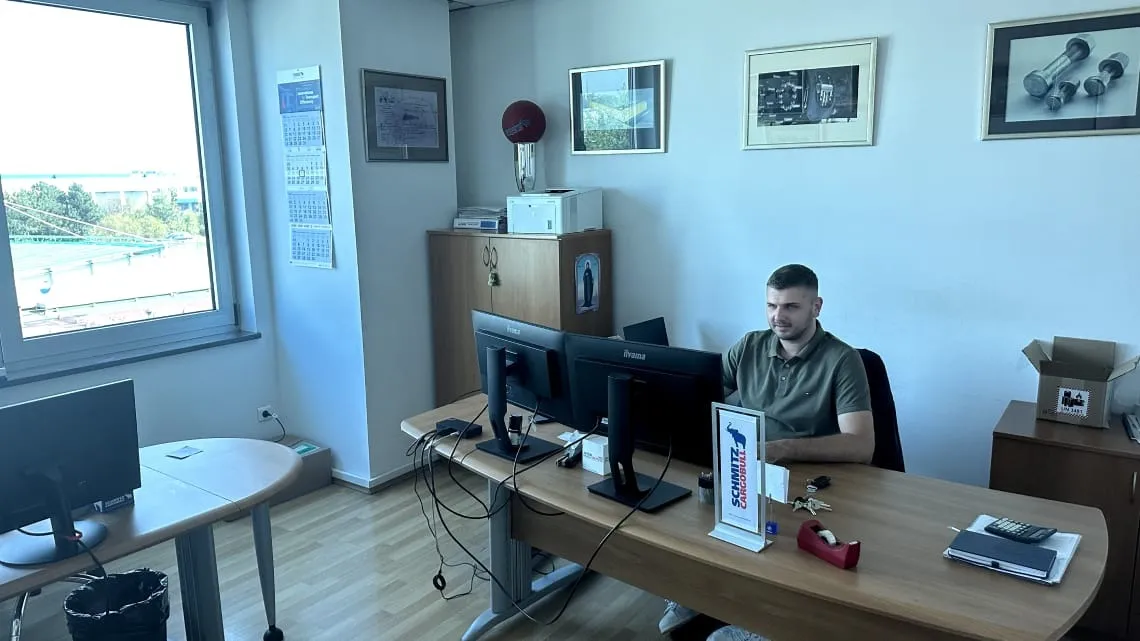 A friendly Schmitz Cargobull team member working at a bright office desk with dual monitors.