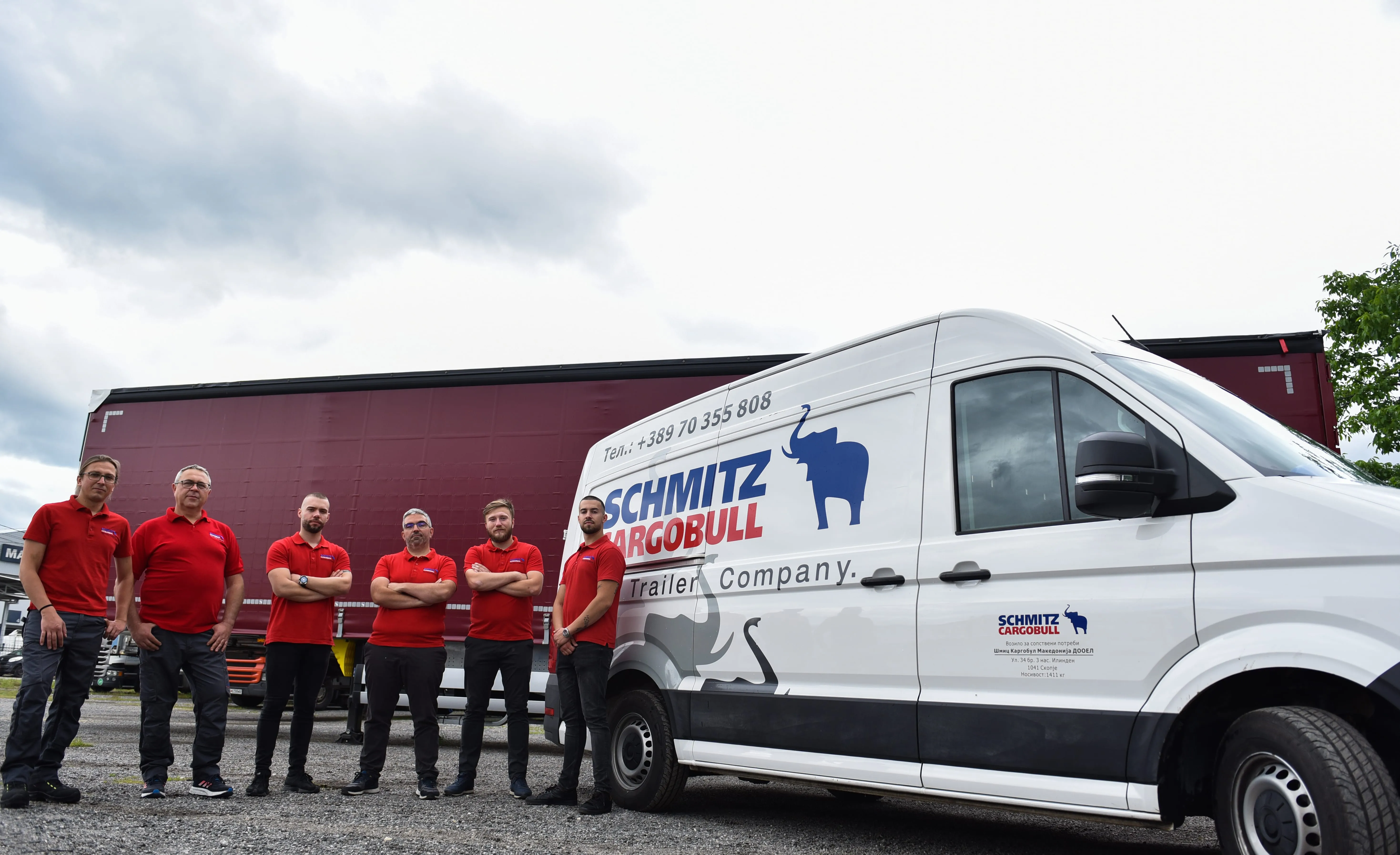 A group of Schmitz Cargobull employees in red shirts stand proudly by a branded company van and trailer.