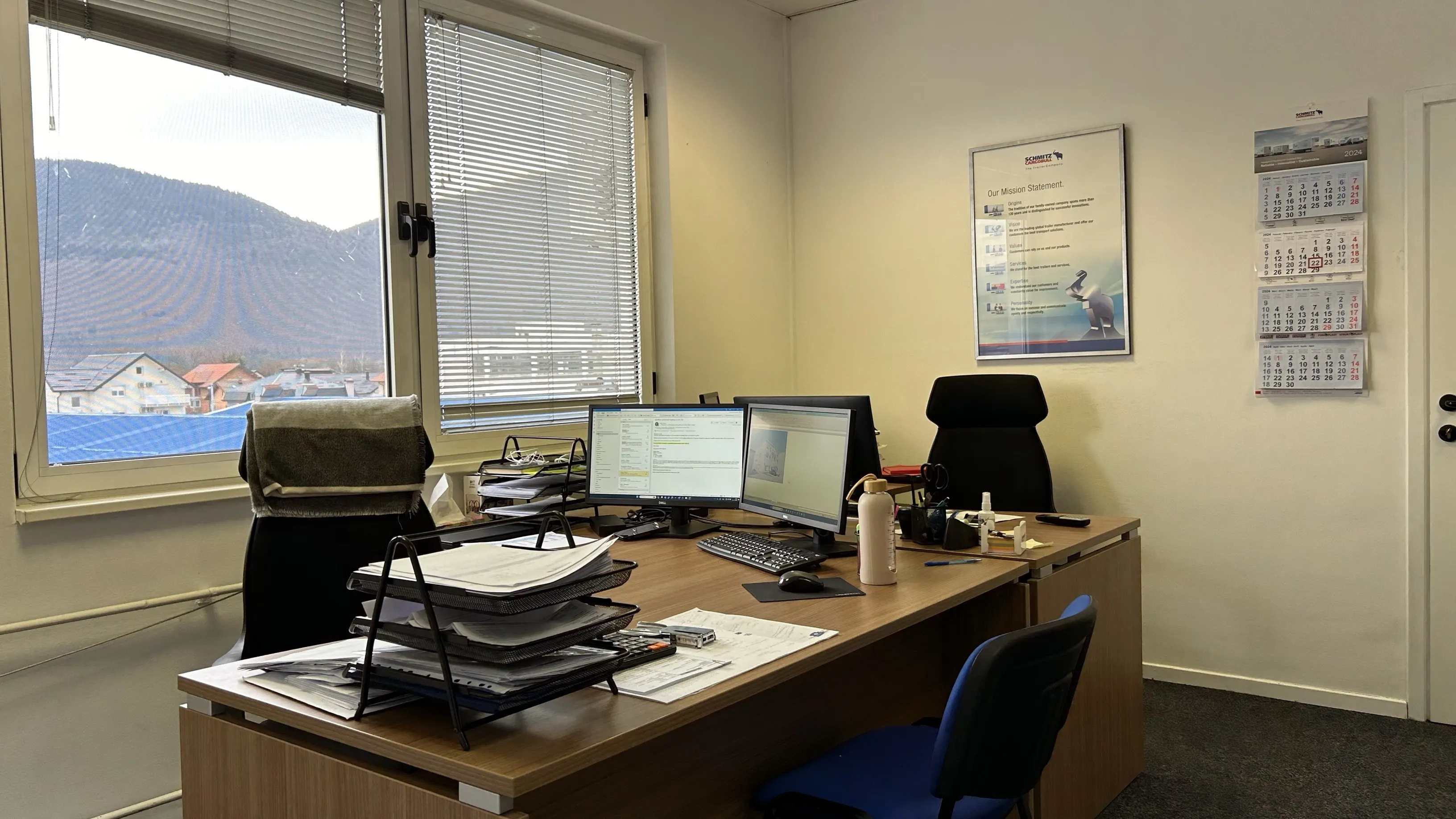 Cozy office at Schmitz Cargobull AG with dual monitors, lots of paperwork, and a stunning mountain view.