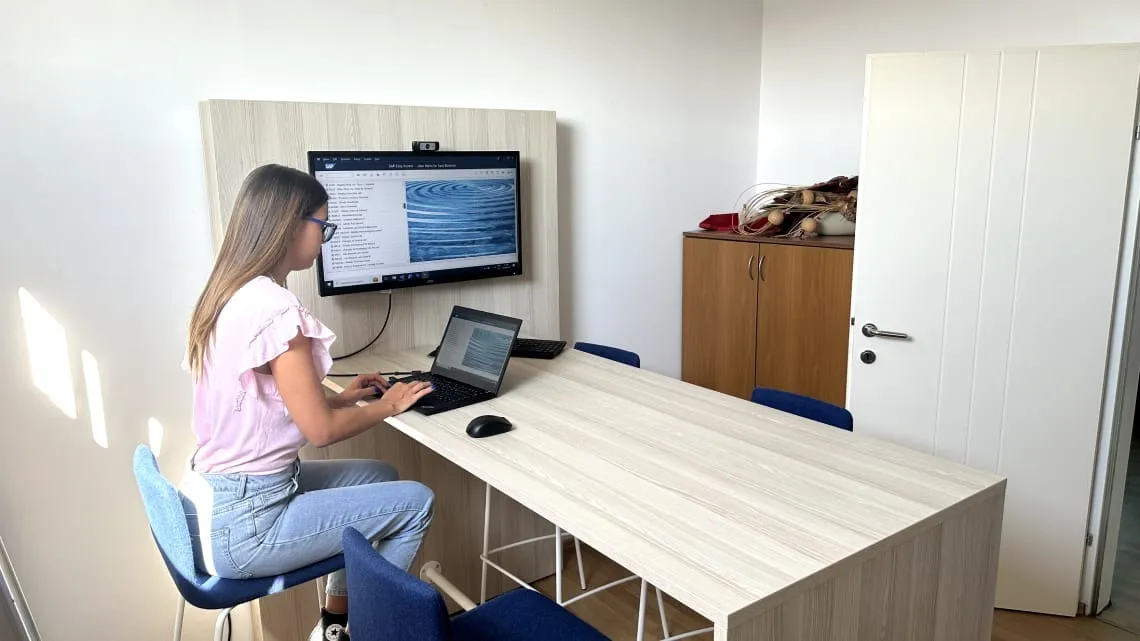 Young woman working on a laptop in a modern office, with a monitor displaying a presentation.